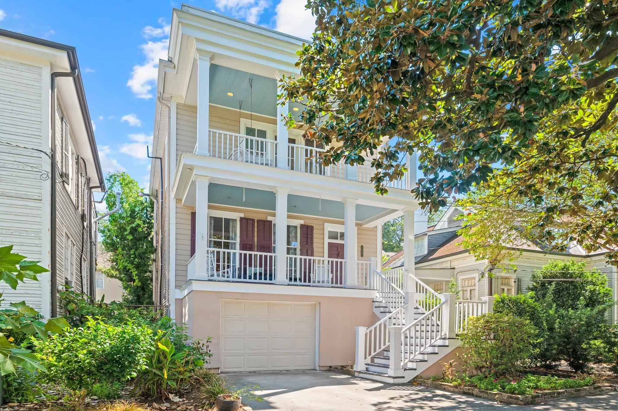A multi-story house with a porch and staircase, surrounded by greenery and trees, with sunny weather and blue sky.