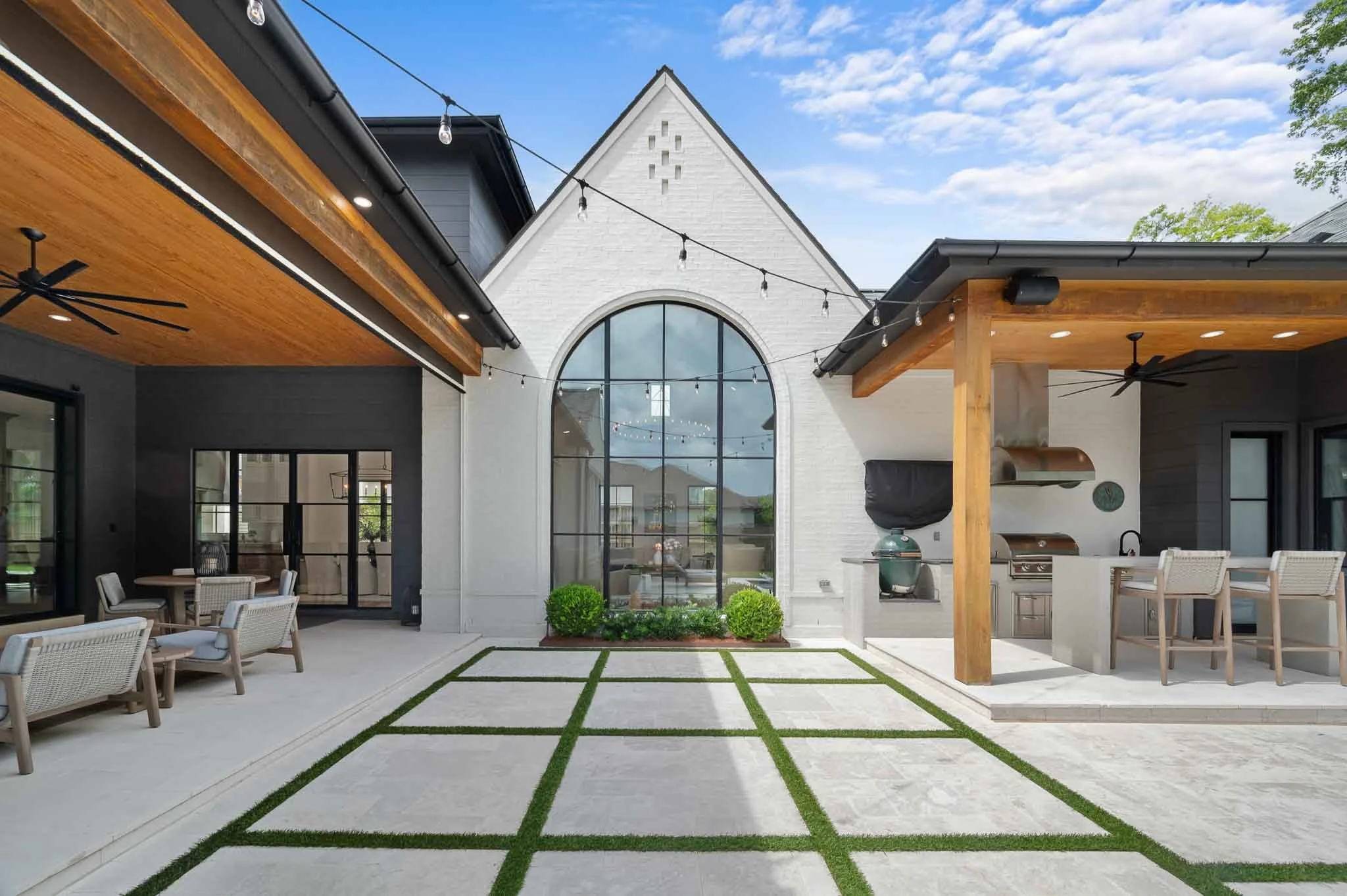 Modern backyard patio with outdoor kitchen, seating area, string lights, and large arched window on a white house with gray and black exterior walls.