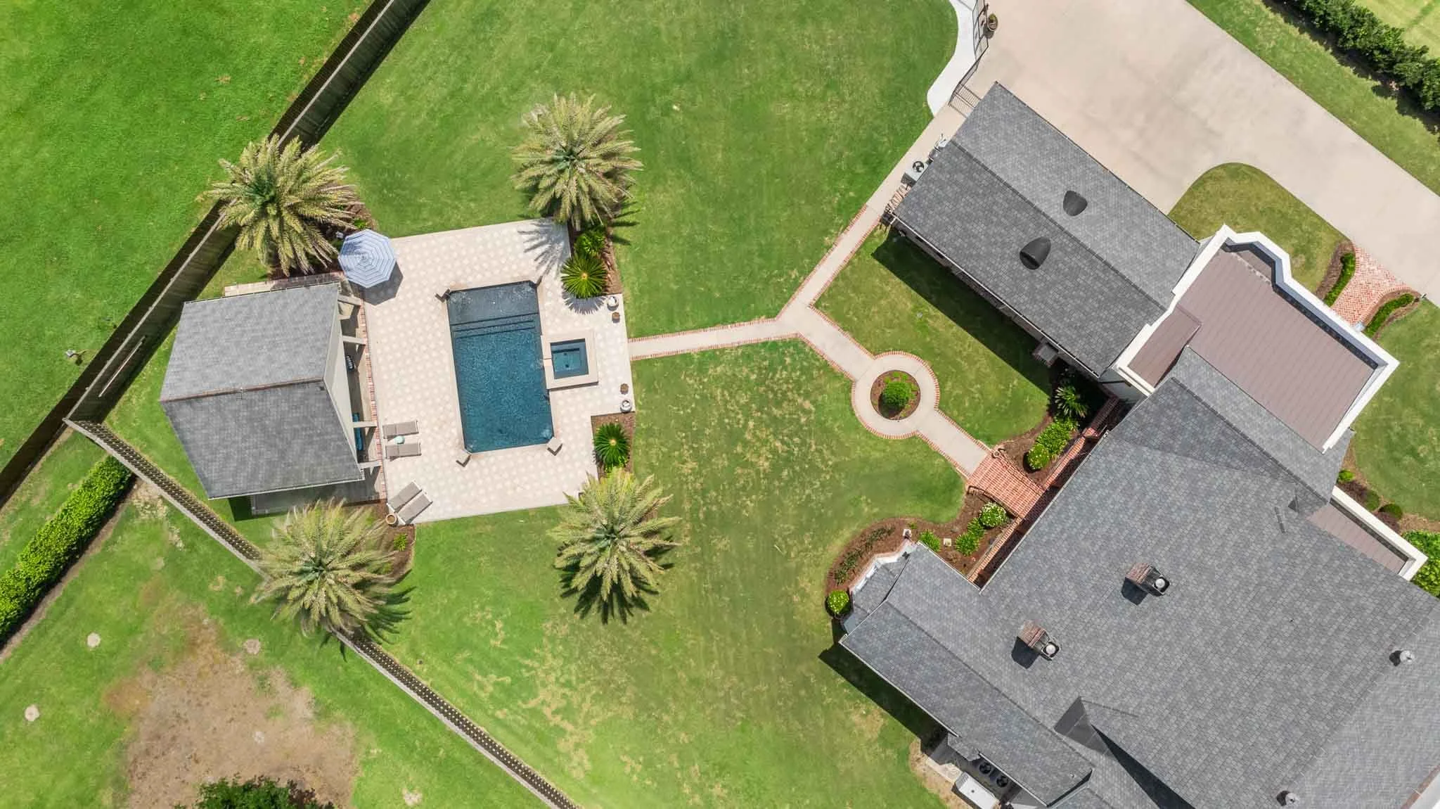 Aerial view of a backyard with a swimming pool, hot tub, palm trees, grassy area, paved pathways, and surrounding houses.