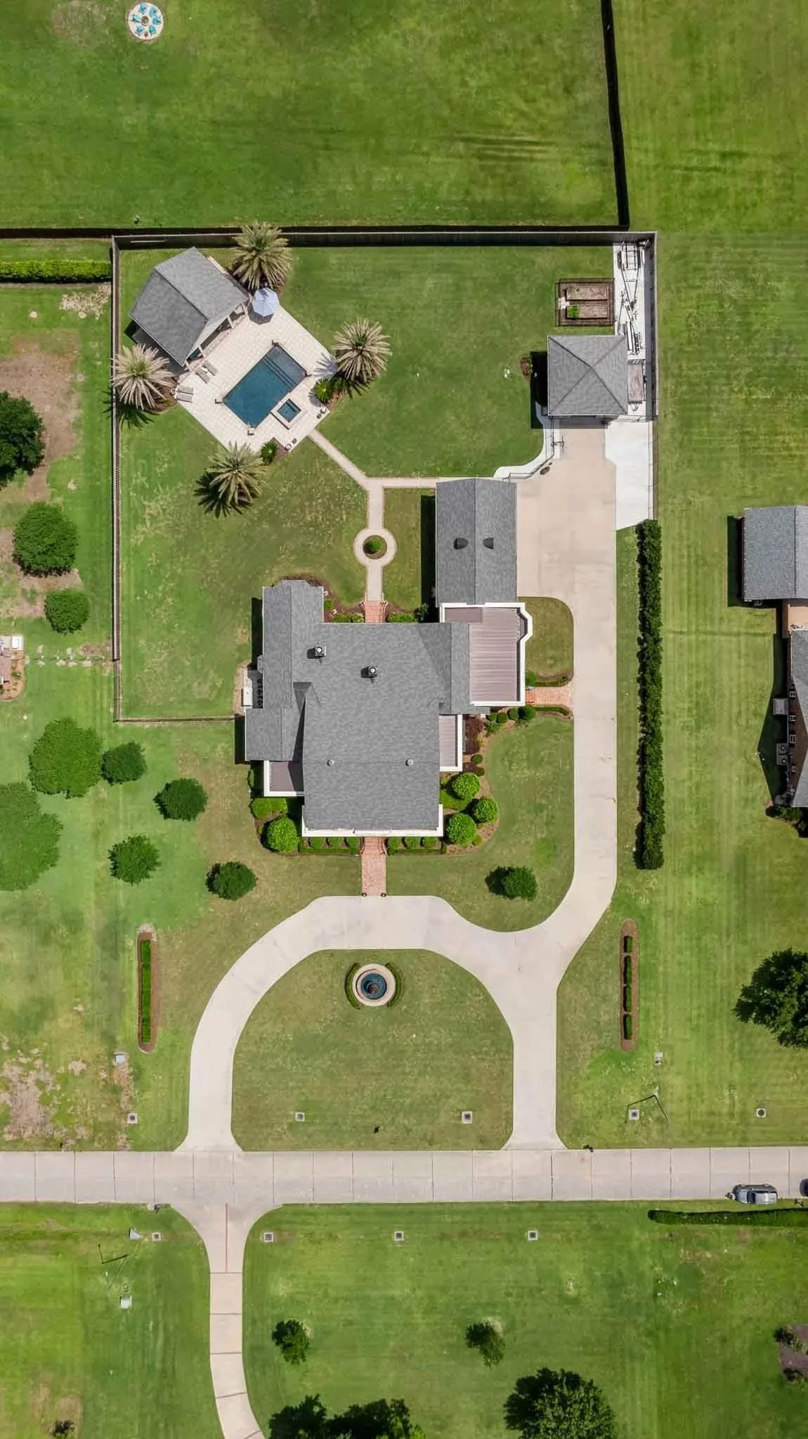 Aerial view of a large house with a gray roof, a front walkway, a circular fountain in the front yard, a driveway, a backyard with a pool, palm trees, and neatly landscaped lawns.