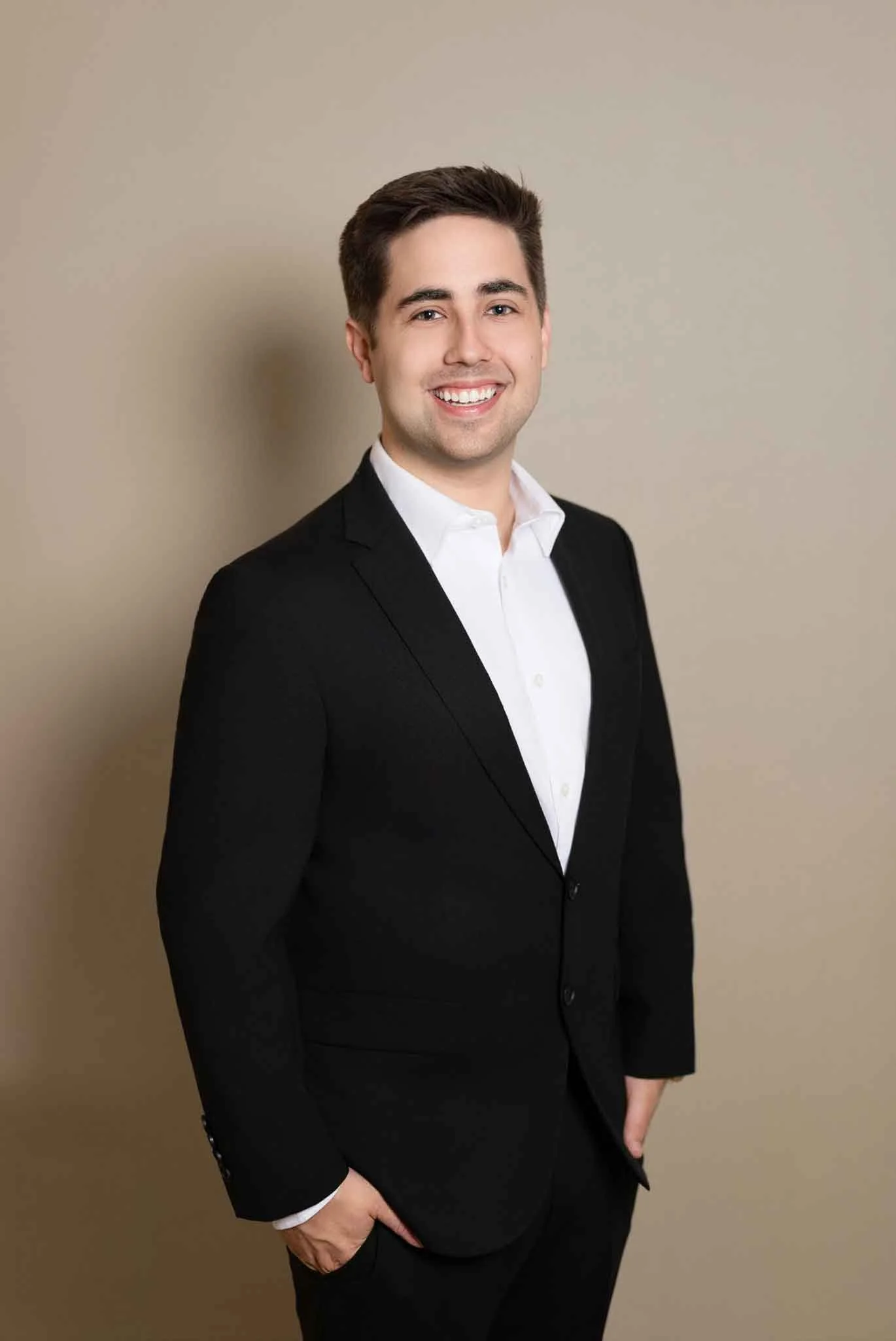 Portrait of a young man in a black suit and white shirt standing against a plain beige background, smiling with hands in pockets.