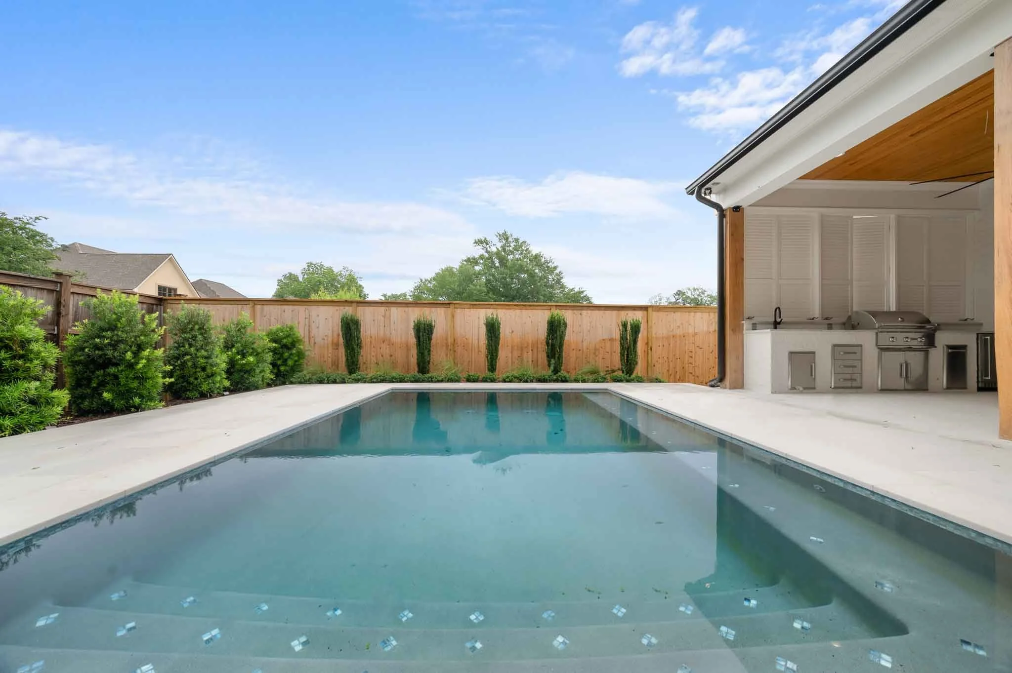 Backyard swimming pool with clear water, surrounded by concrete deck, lush green bushes, and a wooden fence with tall, slim trees, with a partly cloudy sky overhead and an outdoor kitchenette with grill on the right.