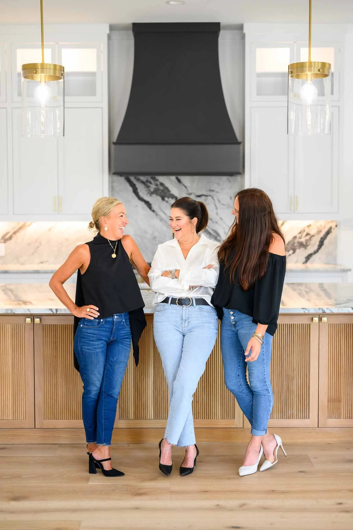 Three women laughing and talking in a modern kitchen with white cabinets, marble backsplash, and black and gold accents.