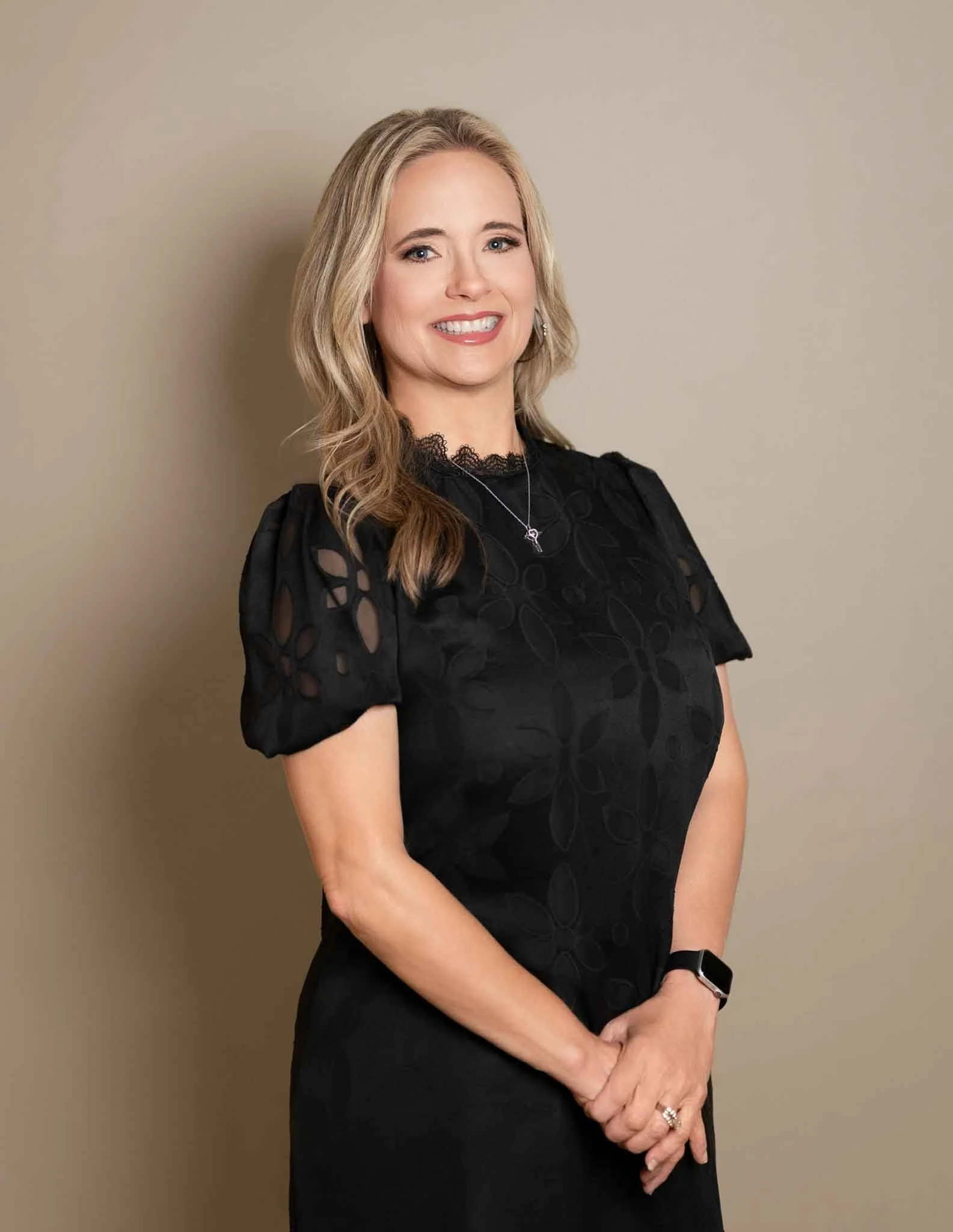 A woman with blonde hair and fair skin smiling, wearing a black dress with cutout floral patterns on the sleeves, a necklace, a watch on her left wrist, and rings on her fingers, standing against a plain beige wall.