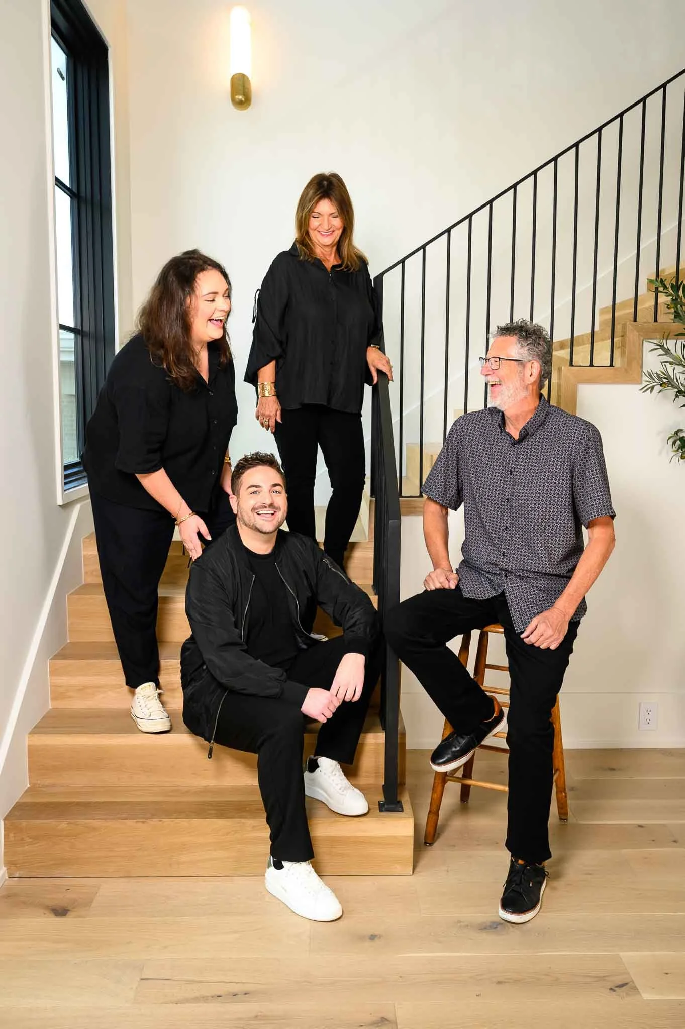 Four people, two women and two men, are gathered on a staircase indoors, laughing and conversing. One woman is standing at the top of the stairs, another is leaning on the railing, one man is sitting on the stairs, and the other man is standing with a foot resting on the bottom rung, engaging in conversation. They are dressed casually.