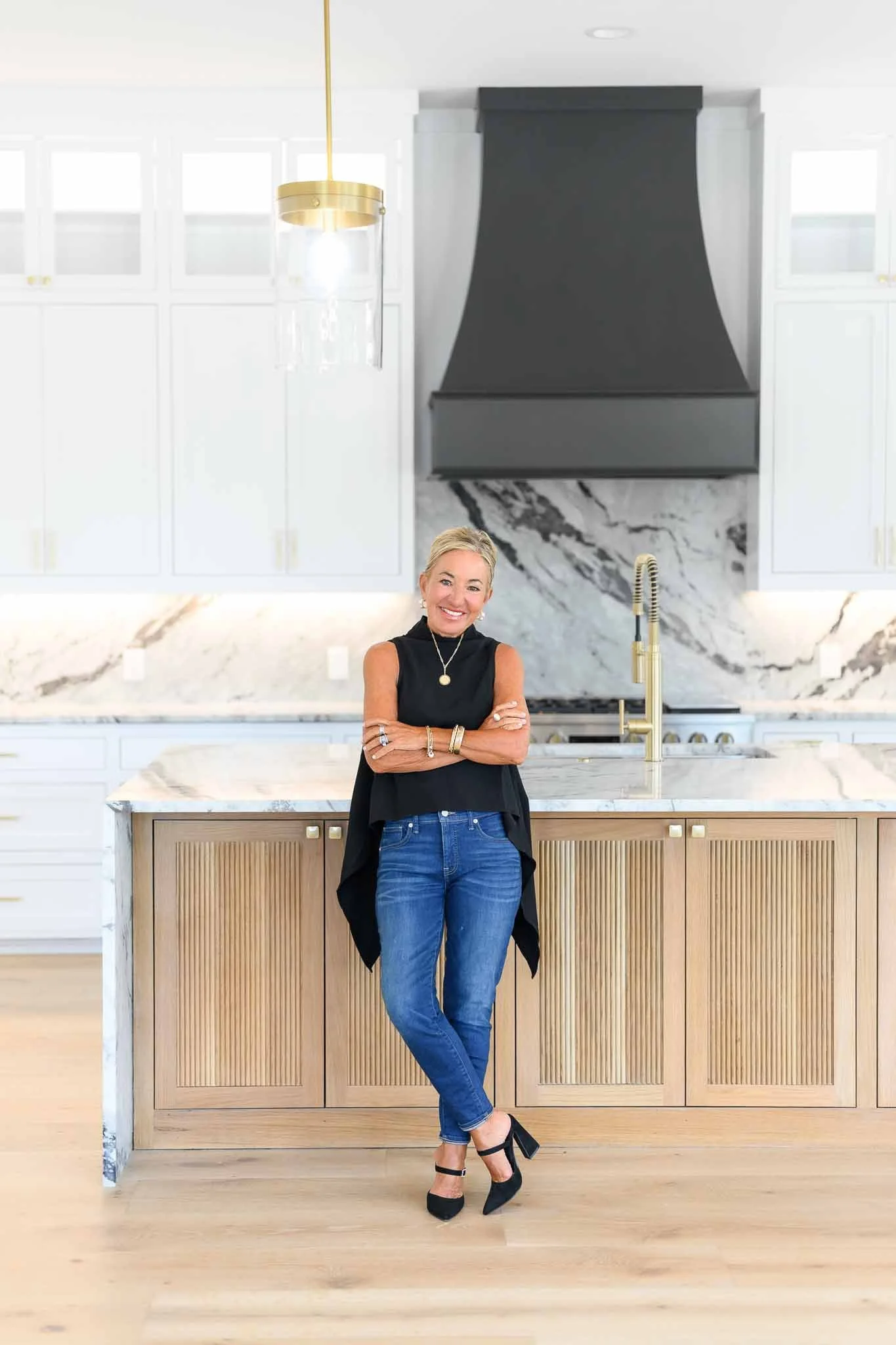 A woman with blonde hair, wearing a black sleeveless top, blue jeans, and black high heels, standing in a modern kitchen with a marble island, gold accents, white cabinetry, black range hood, and gold faucet, smiling at the camera.