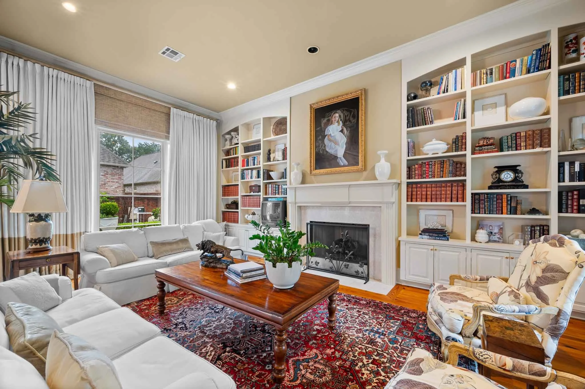 A cozy living room with a white sofa, a wooden coffee table, and a patterned armchair. There's a fireplace with a framed portrait above it, and built-in bookshelves filled with books and decorative items. Large windows with white curtains let in natural light.