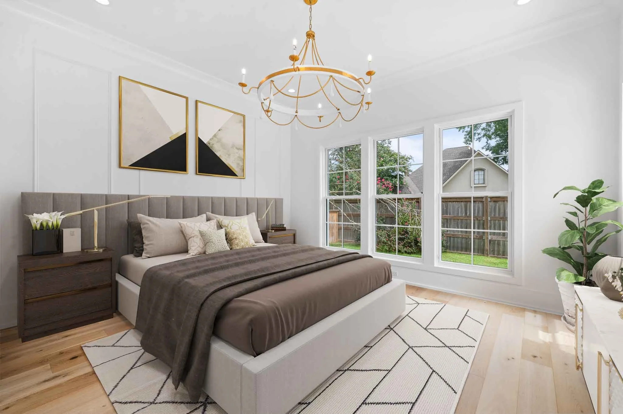 Bright bedroom with large windows, beige bed, gray bedcover, multiple pillows, wooden nightstands, modern artwork, chandelier, patterned rug, and green plant.