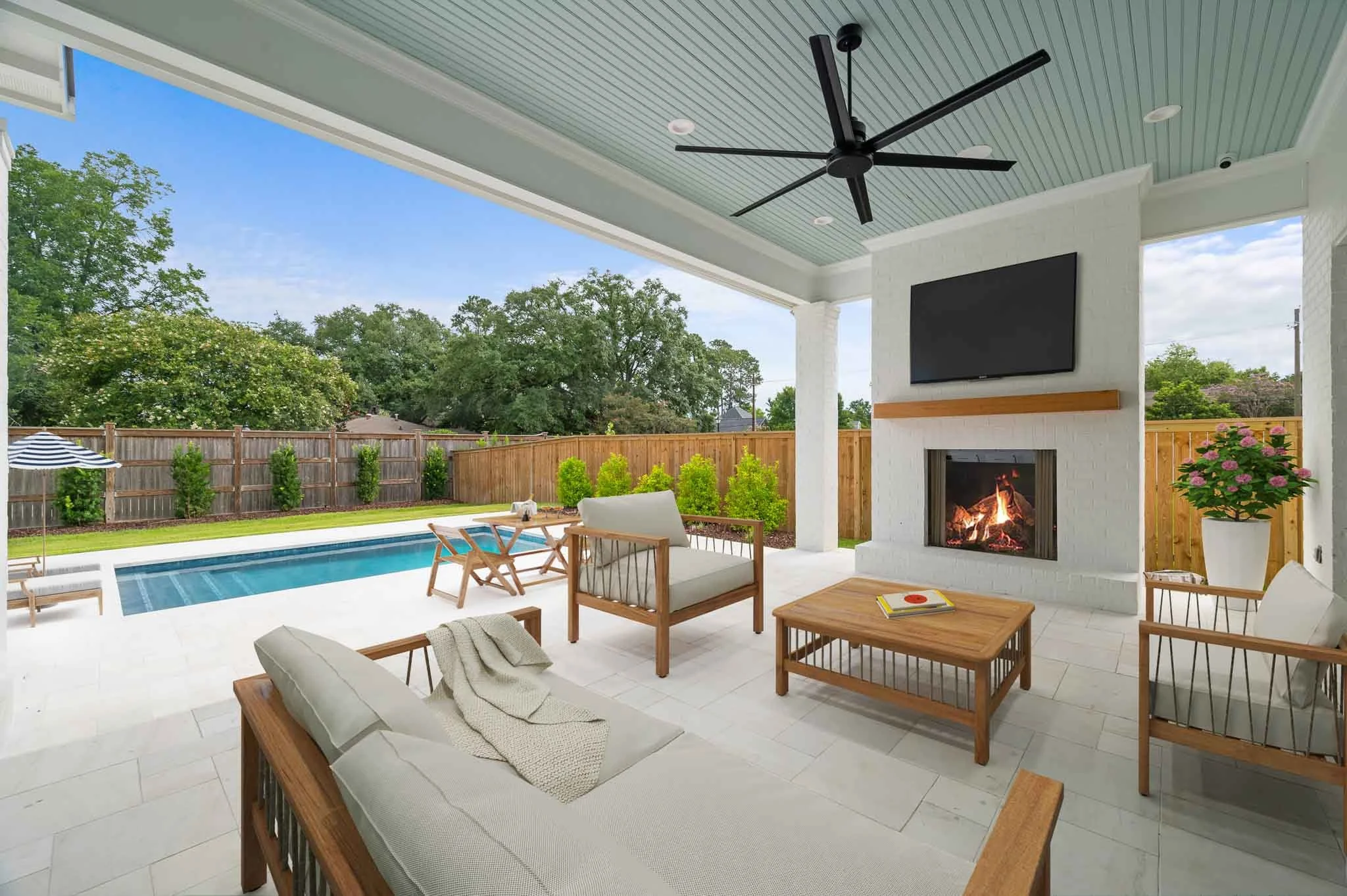 Covered patio with outdoor furniture, a fireplace, a television, and a view of a backyard with a swimming pool, green grass, trees, and a wooden fence.