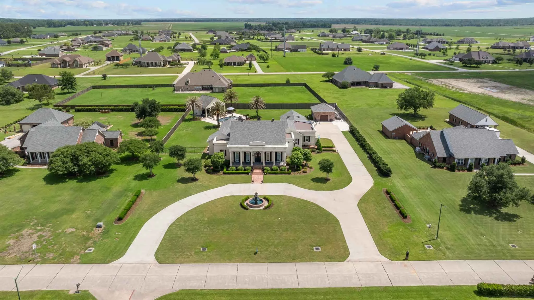 Aerial view of a suburban neighborhood with large, well-maintained houses, green lawns, and trees. The central house features a fountain in its front yard. Roads and driveways are visible, with open fields in the background.