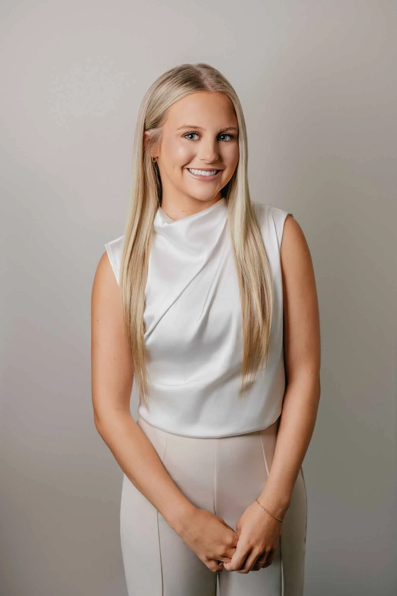 A young woman with long blonde hair, wearing a white sleeveless top and beige pants, smiling and standing against a neutral background.