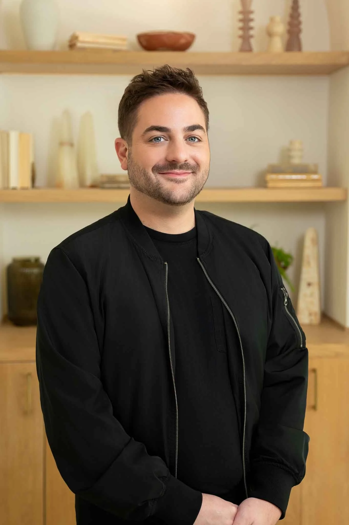 A young man with light skin, short dark hair, and a beard, smiling and wearing a black jacket, standing indoors in front of a wooden shelf with decor and books.