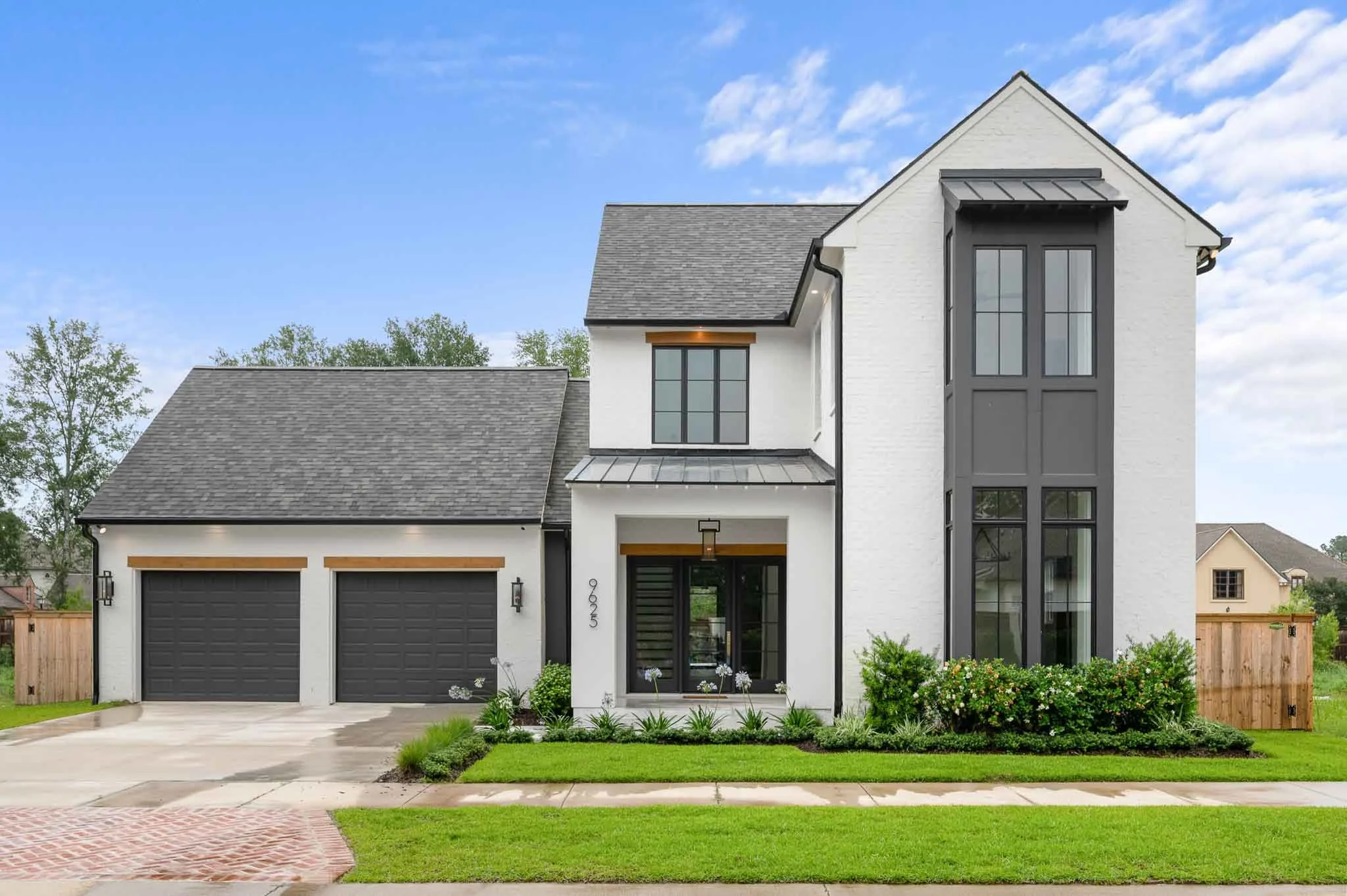 Modern two-story white house with black trim, gray roof, and black garage doors, surrounded by green lawn and landscaped garden.