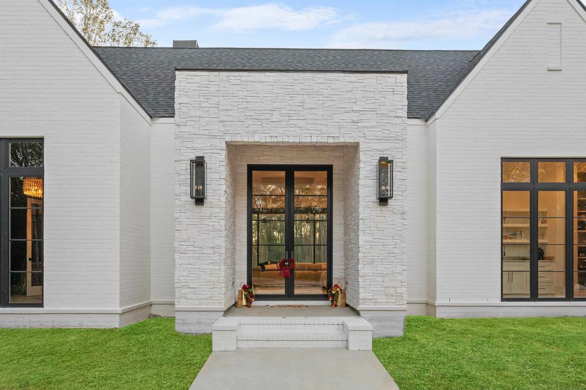 Front entrance of a modern white house with black double doors, decorated for Christmas with wreaths and poinsettias, staircase leading up to the door, green lawn, large windows, and black outdoor wall lights.