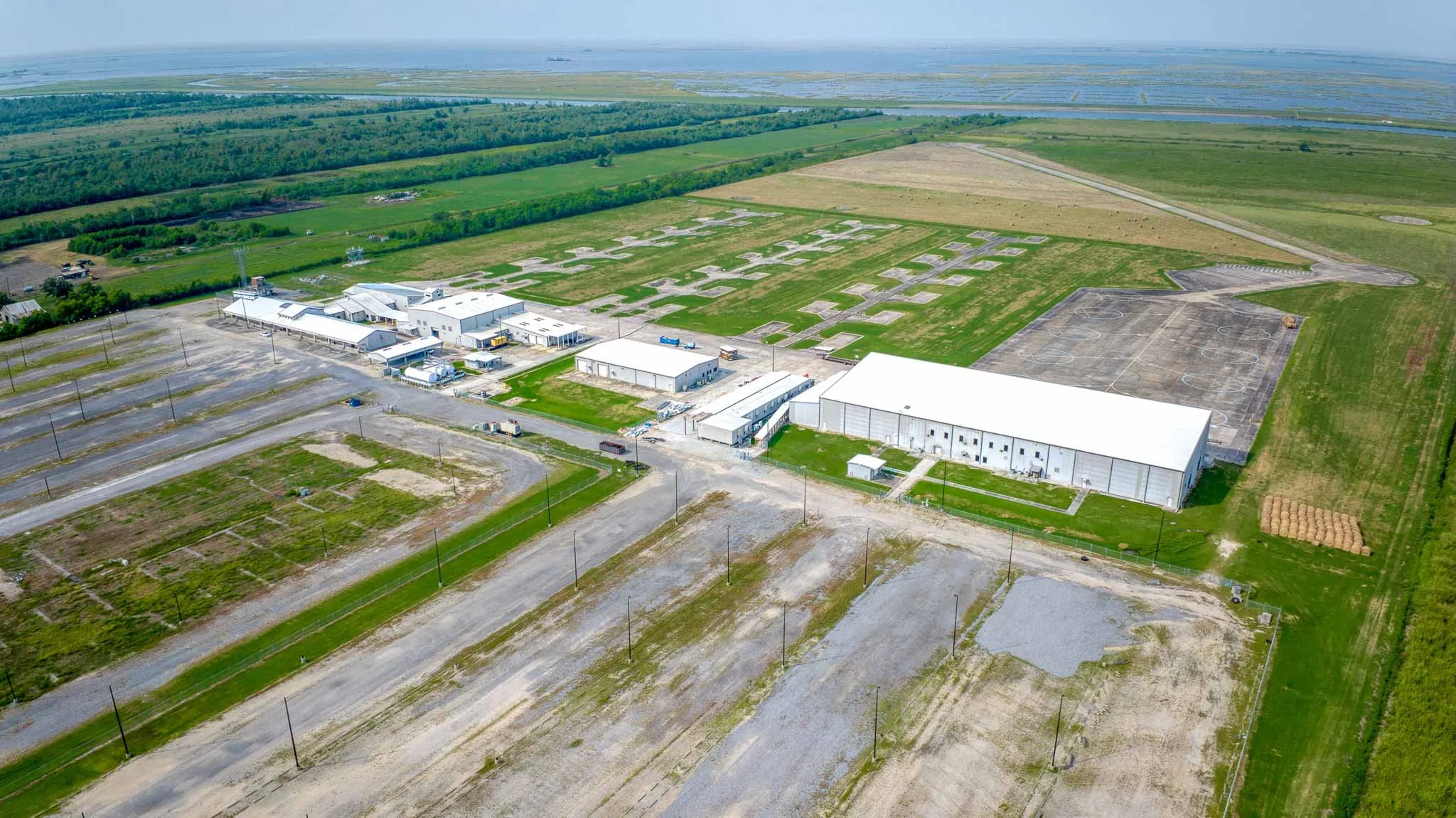 Aerial view of a large indoor sports facility with multiple tennis courts, surrounded by parking lots, green fields, and farmland with ponds in the distance.
