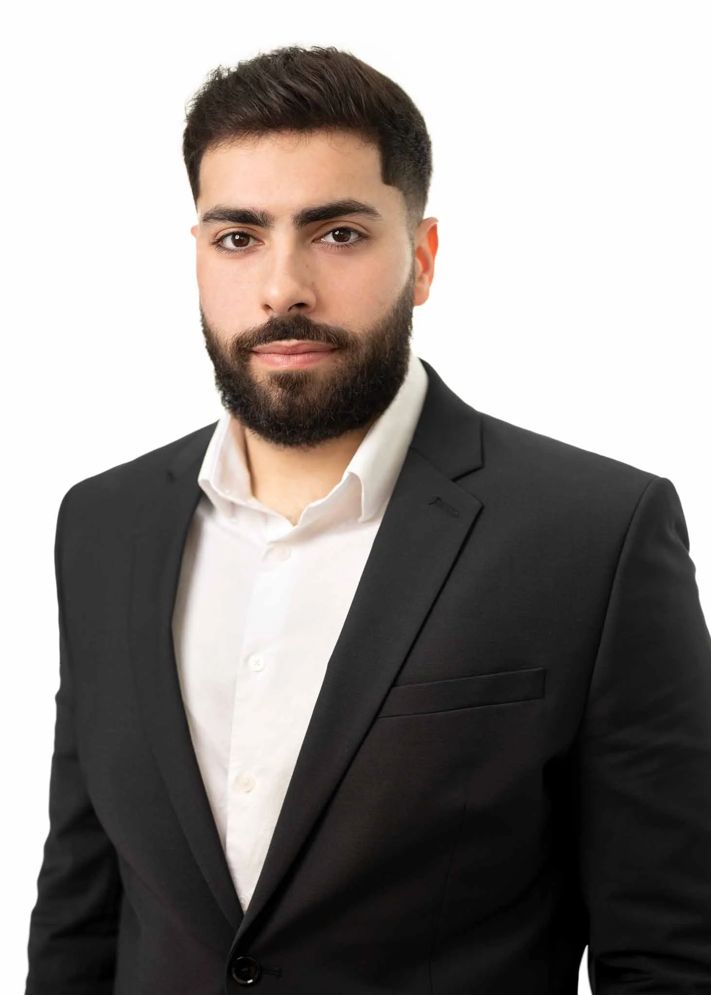 A young man with dark hair and a beard wearing a black suit jacket and a white dress shirt against a white background.