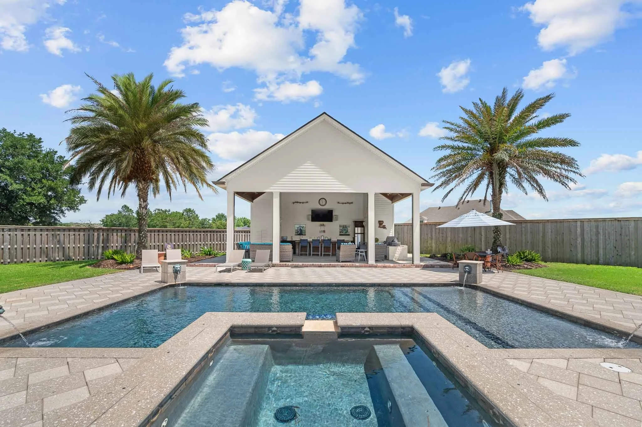 Backyard with a swimming pool and a pool house. The pool has water features and an attached hot tub. The pool house has an outdoor bar, television, and seating area, with two palm trees and a fenced yard.