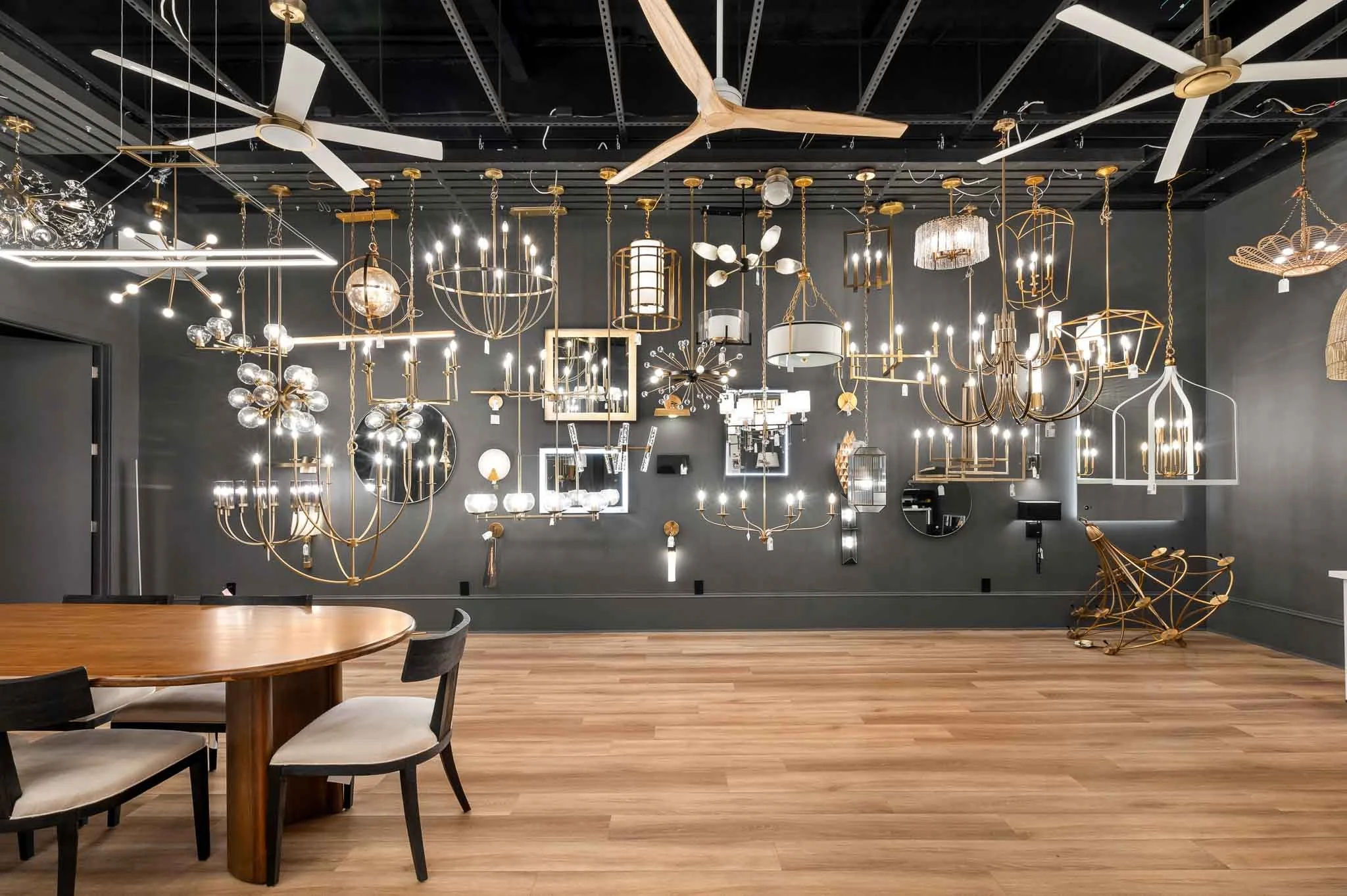 Showroom with various modern light fixtures and chandeliers hanging from a dark ceiling, with a wooden table and chairs in the foreground.