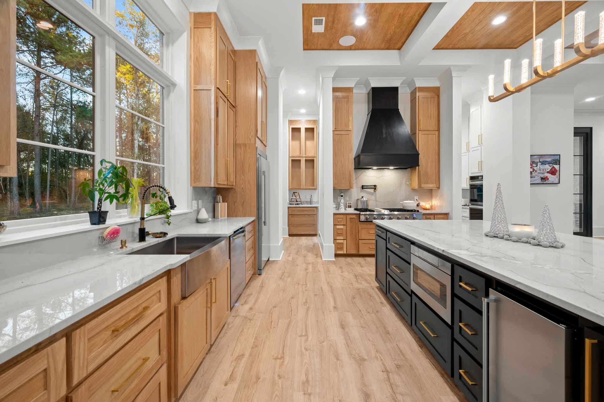 Modern kitchen with large windows, wooden cabinets, black range hood, island with marble countertop, and holiday decorations.