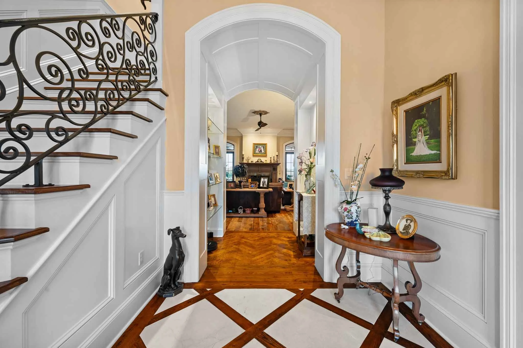 Entryway of a house with a wooden floor, a small wooden console table with decorative items, a gold-framed portrait on the wall, and a staircase with ornate black iron railing on the left. Further into the house, a living room is visible with a fireplace, framed photos, and windows."
