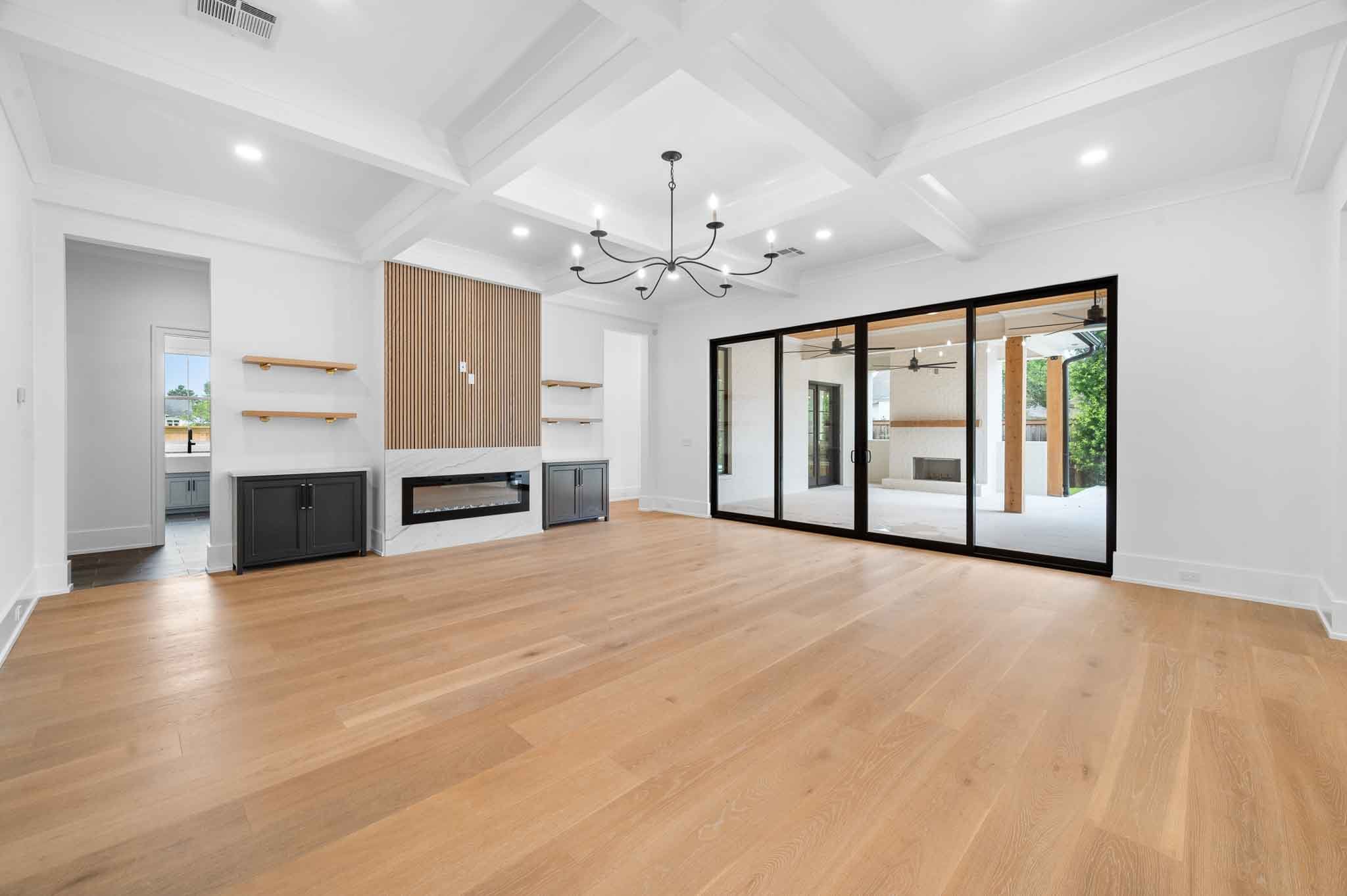 Empty living room with light wood flooring, white walls, a modern black fireplace on a wall with wooden slats, black built-in cabinets, a black chandelier, and large sliding glass doors leading to an outdoor patio area.