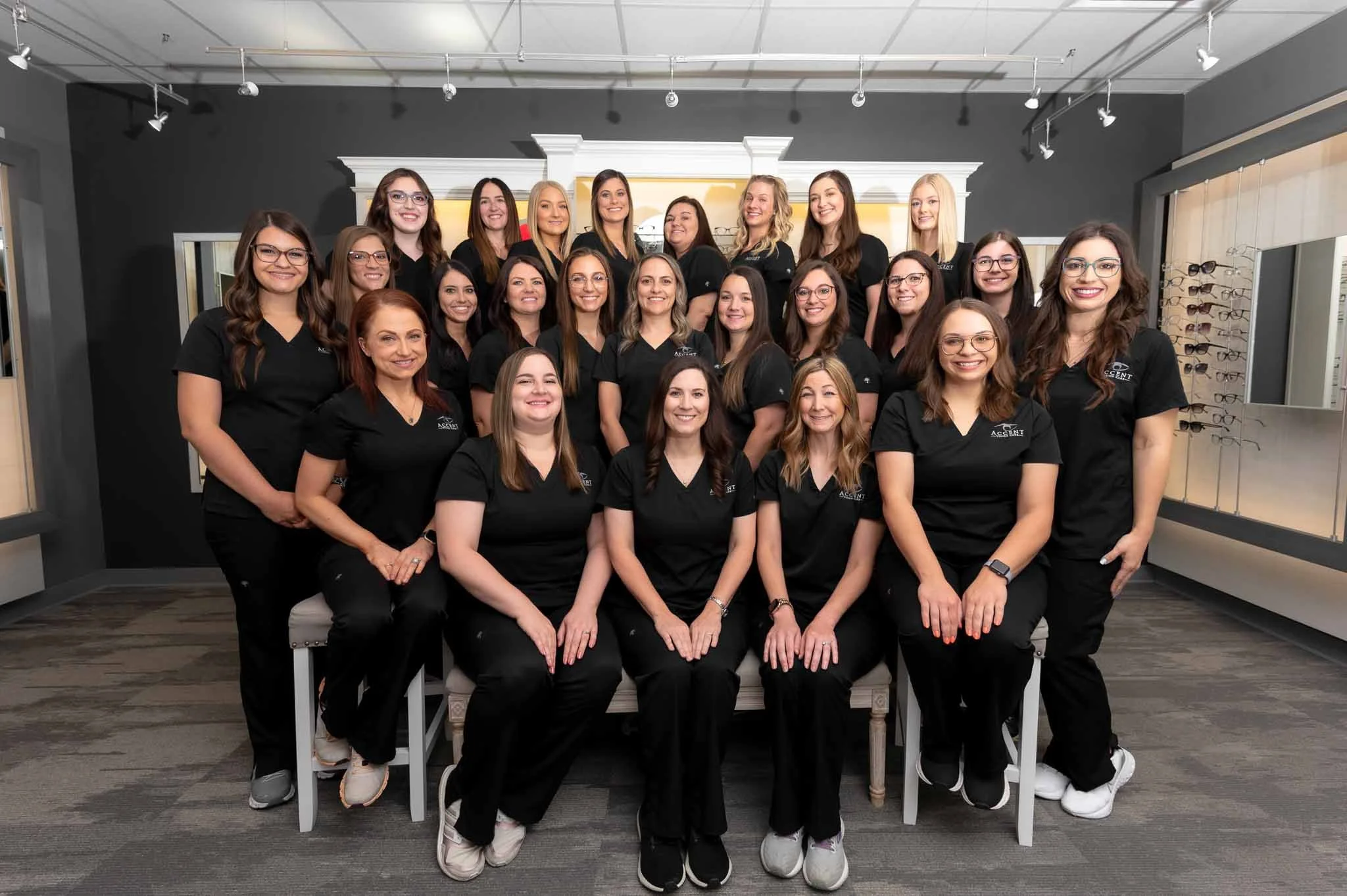 Group of 20 women wearing black scrubs inside an eyewear store, posing for a photo with eyeglasses displayed on the wall behind them.