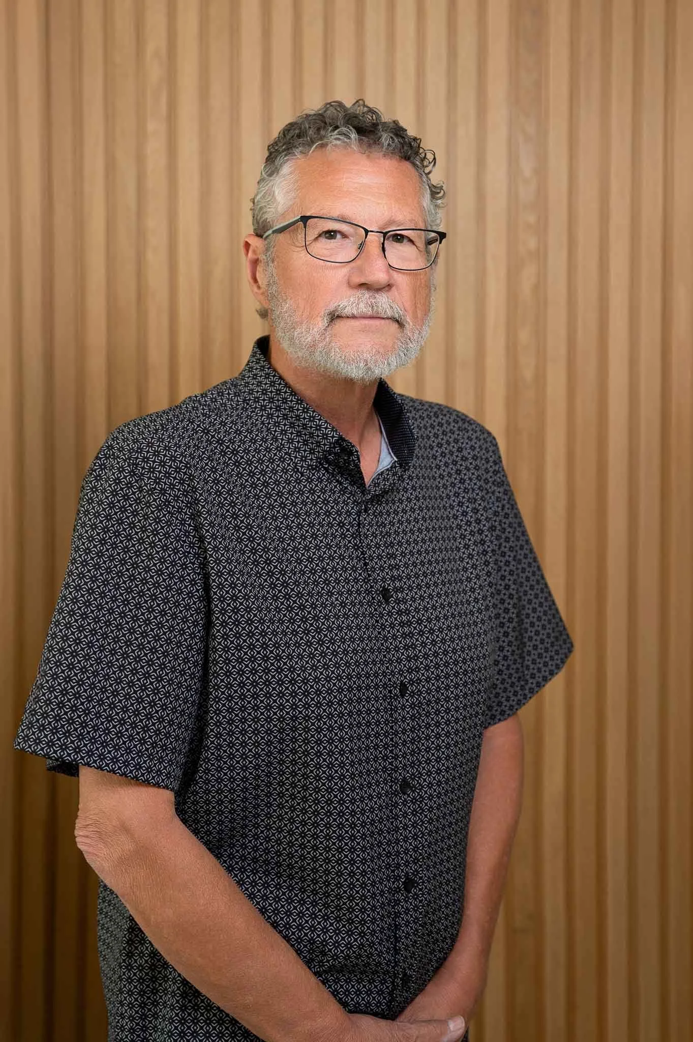 A middle-aged man with gray hair and a beard, wearing glasses and a patterned short-sleeve shirt, standing in front of a wooden-paneled wall.