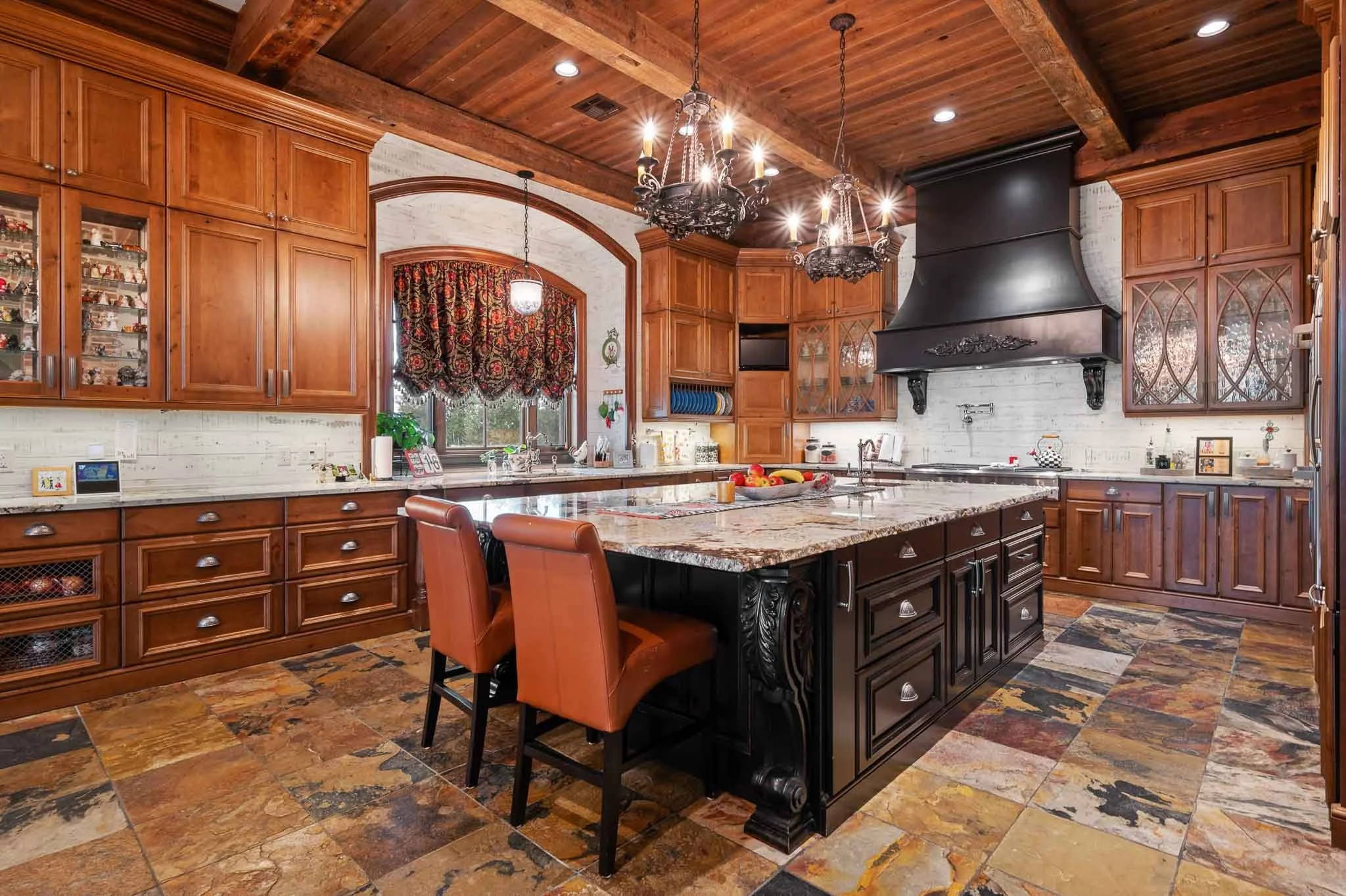 A luxurious kitchen with wooden cabinets, a large central island with a granite countertop, and a black range hood. There are chandeliers hanging from a wooden ceiling, and a window with patterned curtains. The floor is tiled with multicolored stone tiles.