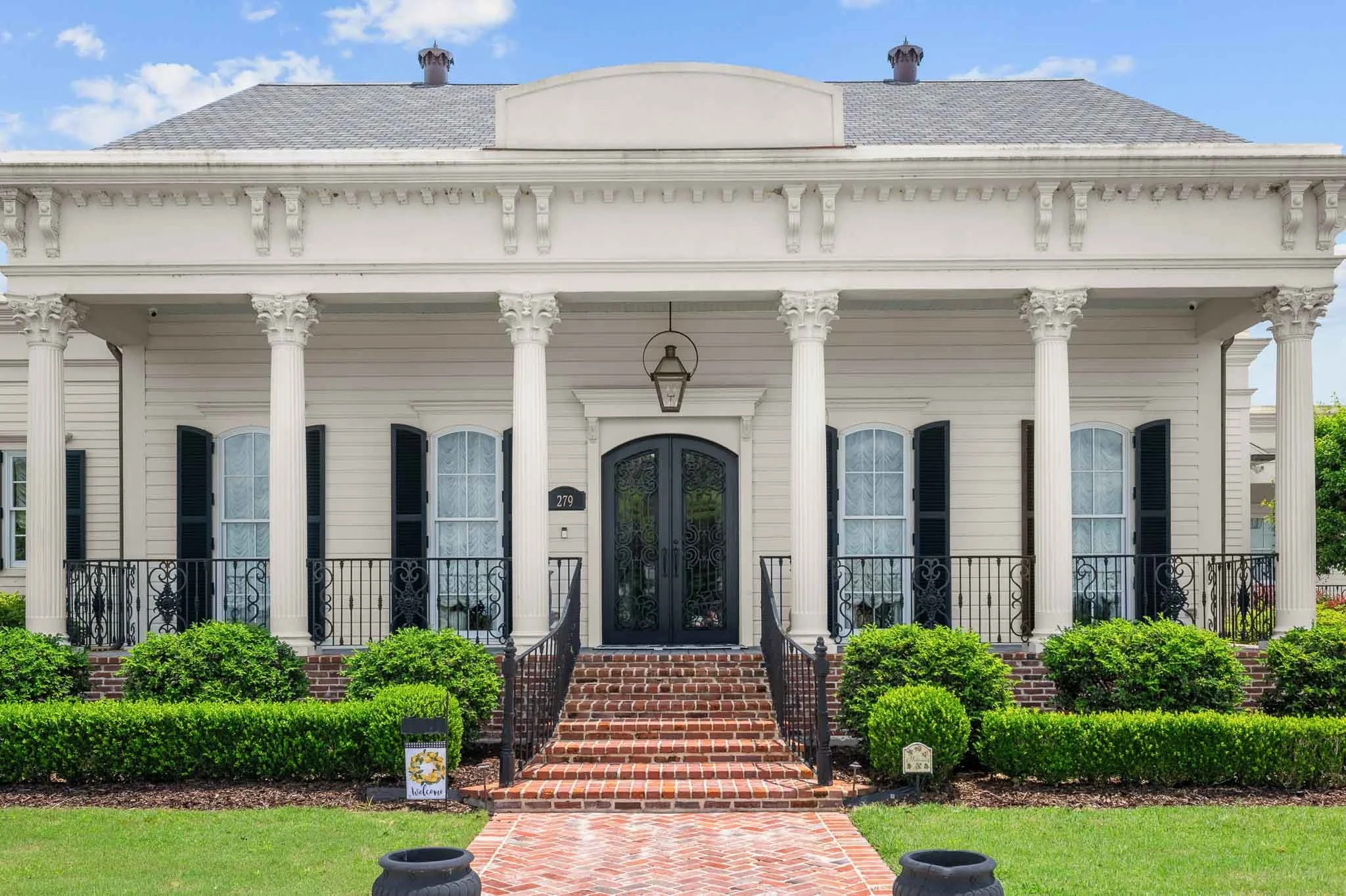 Front view of a white, two-story house with columns, black front door, and brick stairs, surrounded by green bushes and lawn.