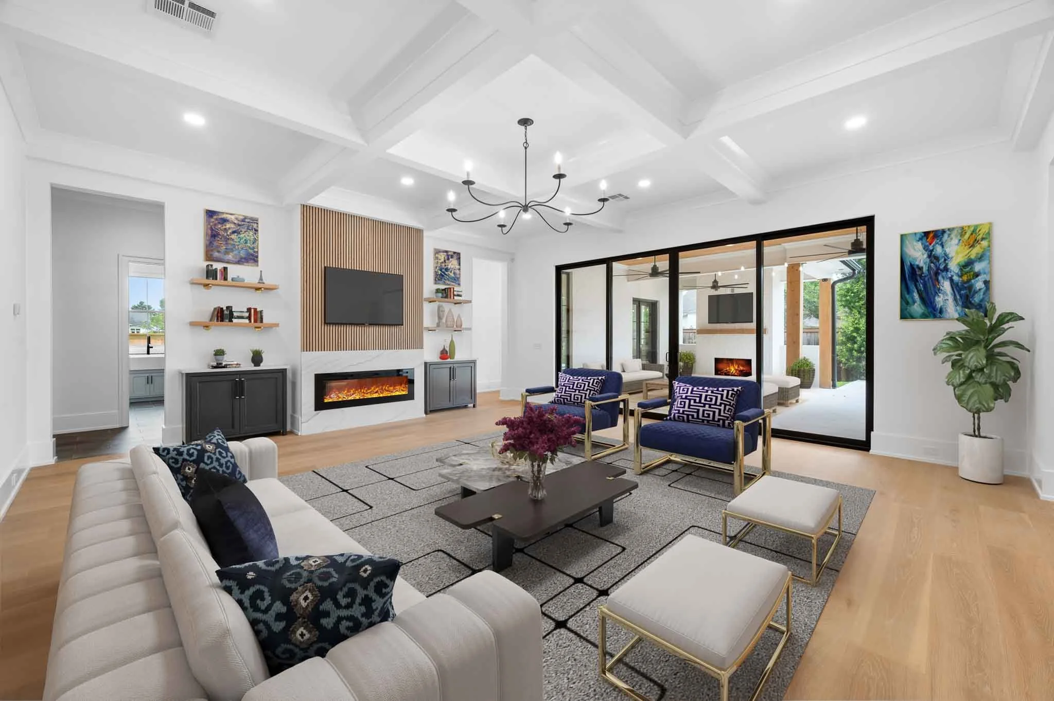 Modern living room with white walls, wooden floors, a white sofa, blue armchairs, and a black coffee table with pink flowers. There is a fireplace, a TV on a wooden panel, and art on the walls. Large glass doors lead to an outdoor patio with seating and ceiling fans.