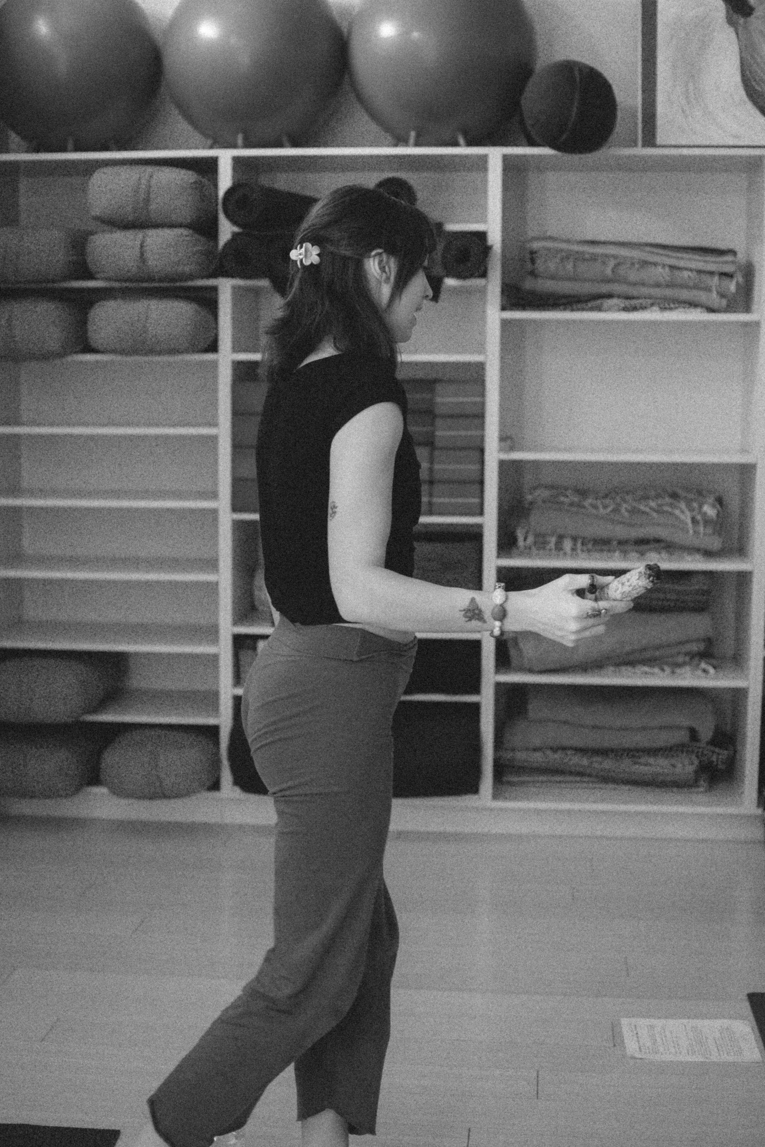 A woman standing in front of shelves filled with foam rollers and yoga mats in a gym or fitness studio, holding a snack and a beverage.
