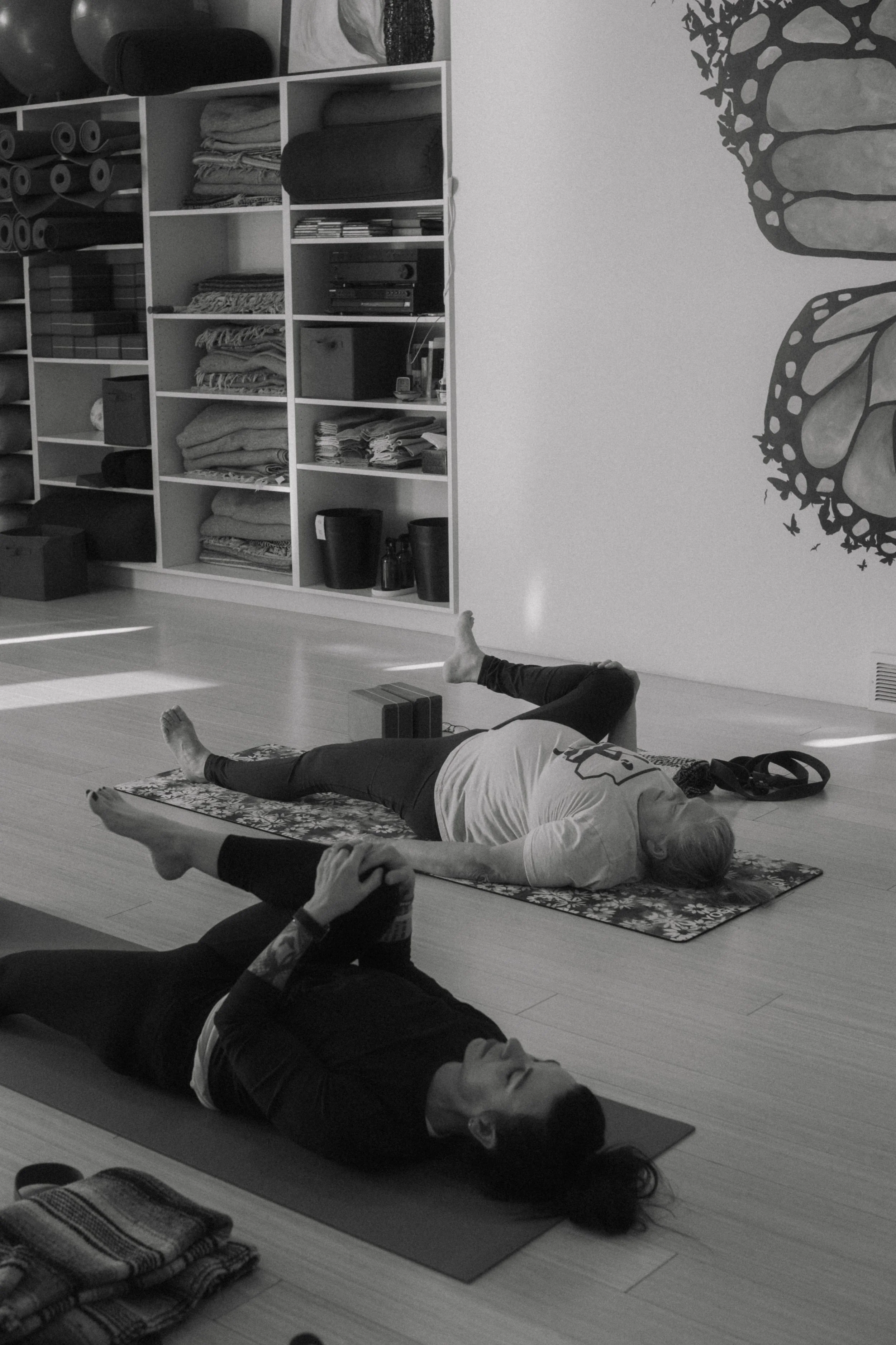 People lying on yoga mats in a yoga class, practicing relaxed poses. The room has shelves filled with folded towels, mats, and some electronic equipment. A gallery wall with artwork or decorative elements is visible.