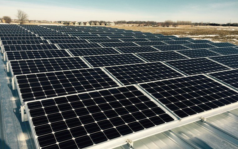 A large field filled with rows of solar panels installed on a rooftop, surrounded by open land with some trees in the distance under a clear sky.