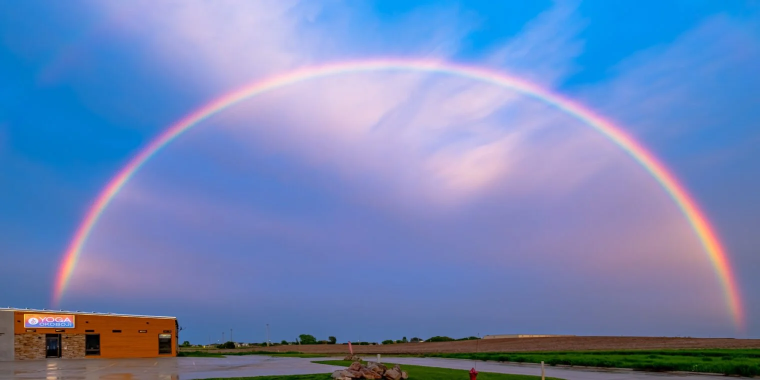 Colorful rainbow arching across a cloudy sky above a rural area with a building labeled 'Yoga Okoboji' and open grassy fields.
