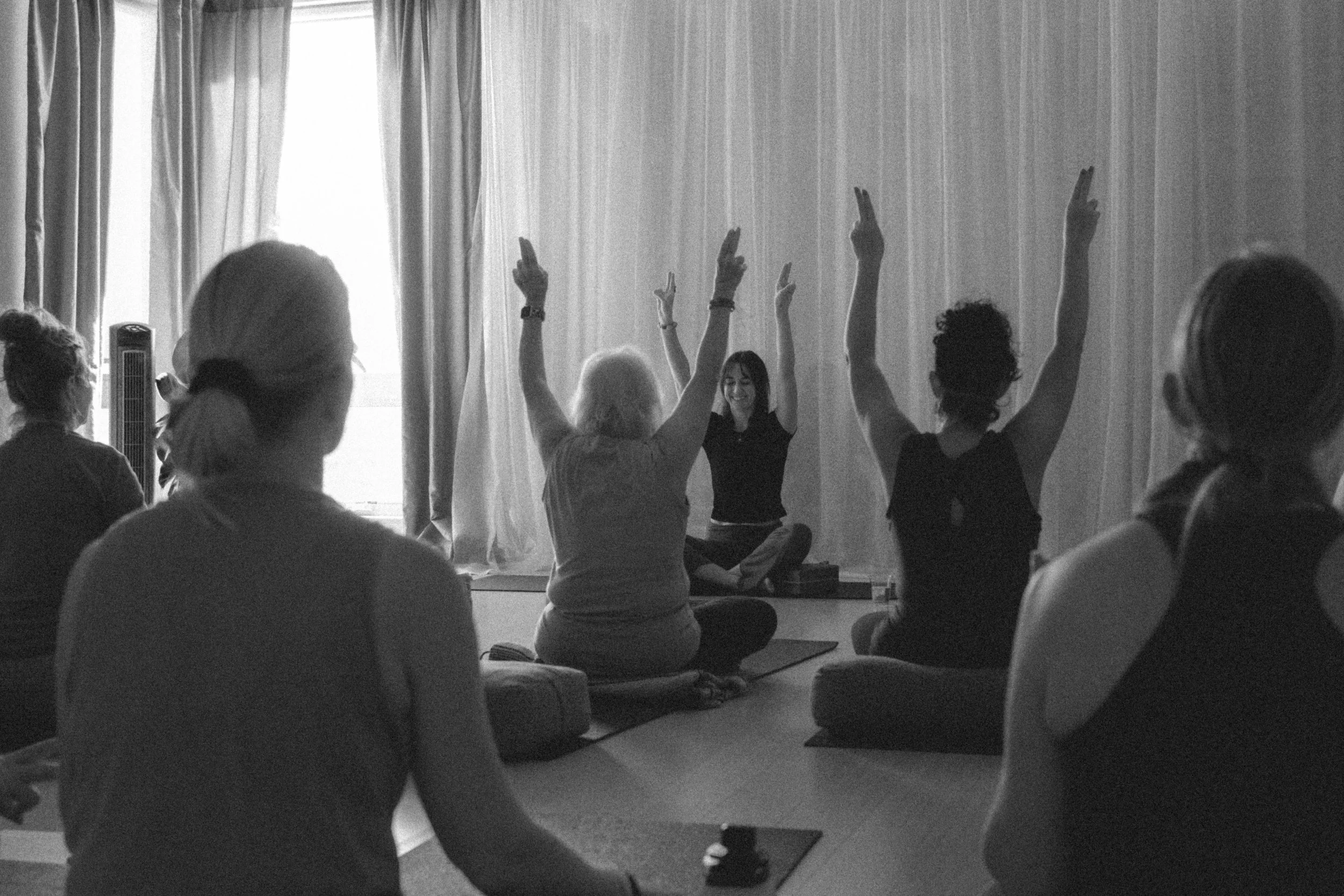 People participating in a yoga or meditation class, sitting cross-legged on mats with some raising their hands in a pose, in a room with curtains and natural light.