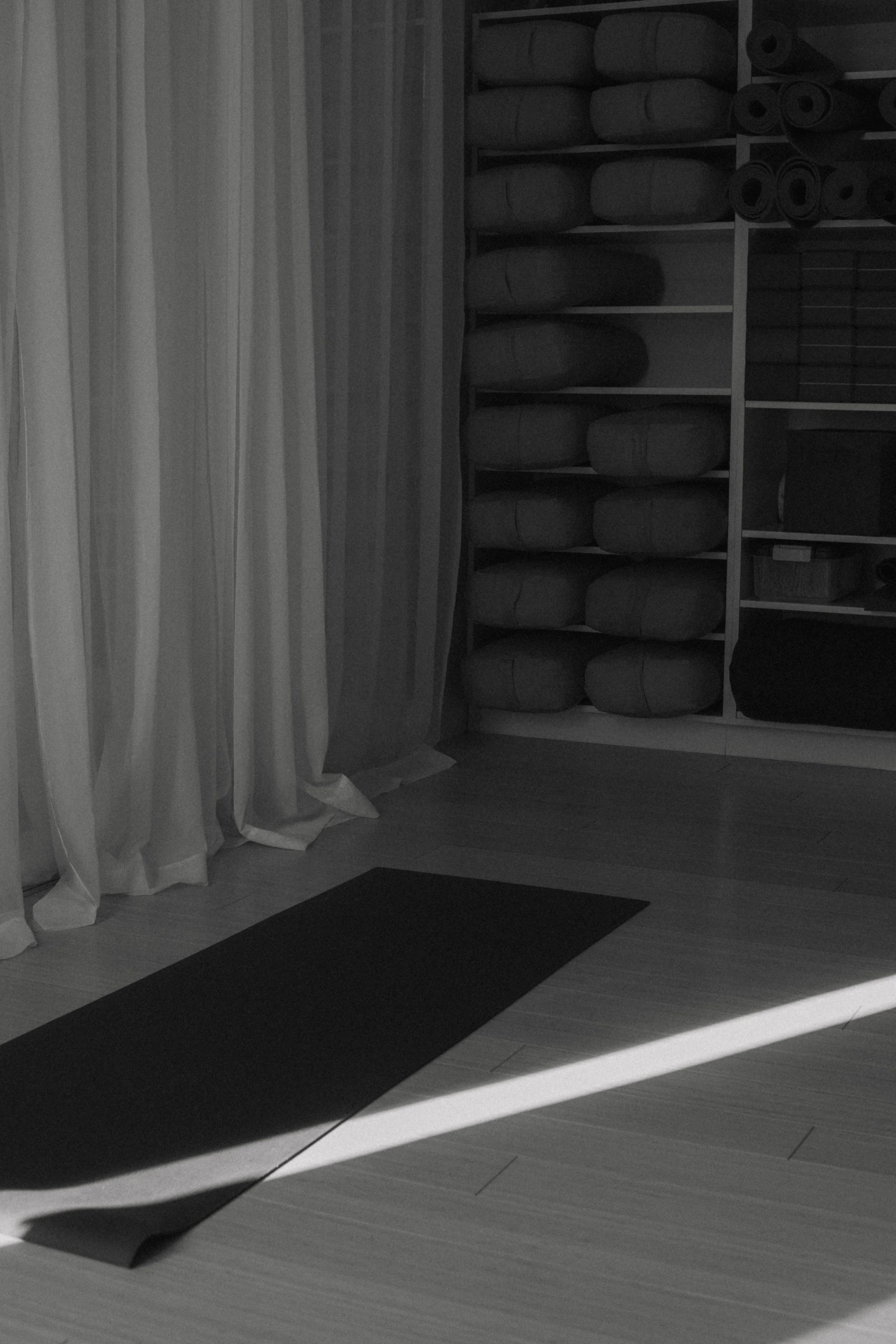 A room with a shelf of rolled-up yoga mats and a mat on the floor, with sunlight creating shadows on the wooden floor and curtains.