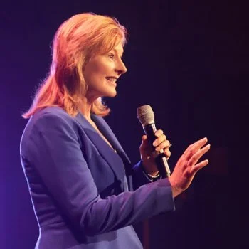 A woman with red hair wearing a blue blazer holding a microphone and speaking on stage.