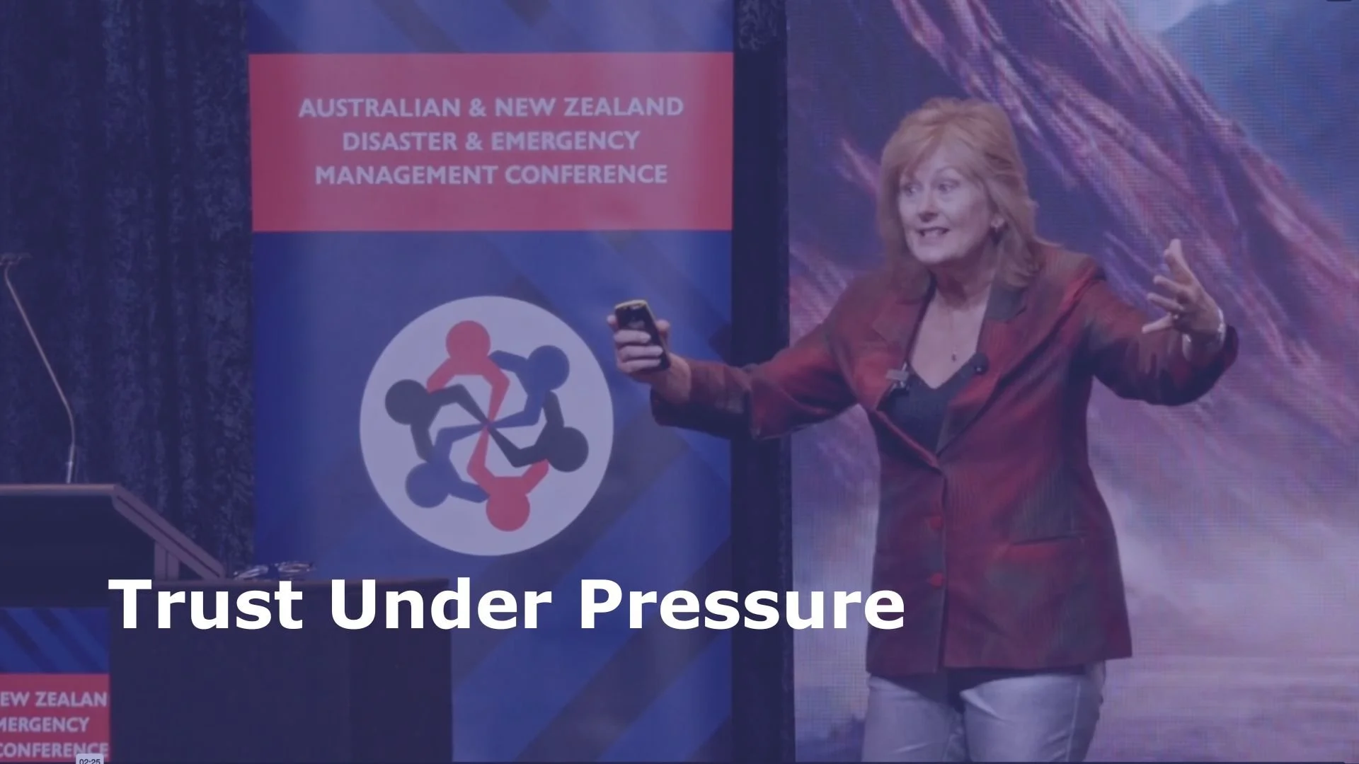 A woman speaking at a conference titled 'Australian & New Zealand Disaster & Emergency Management Conference'. The slide on the background says 'Trust Under Pressure'.