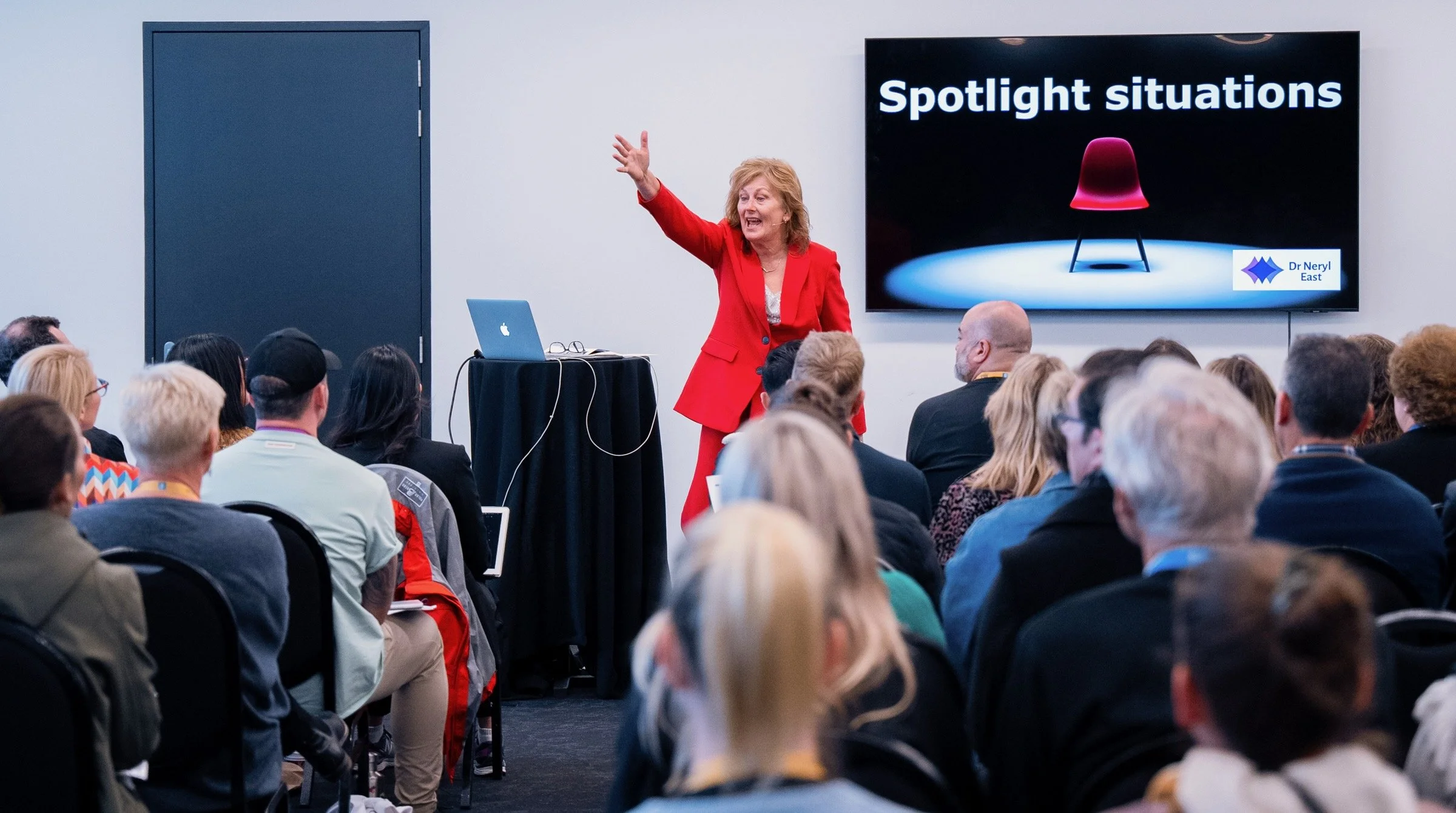 A woman in a red suit giving a presentation in front of a seated audience. There is a large screen displaying the words "Spotlight situations" and an image of a red chair.