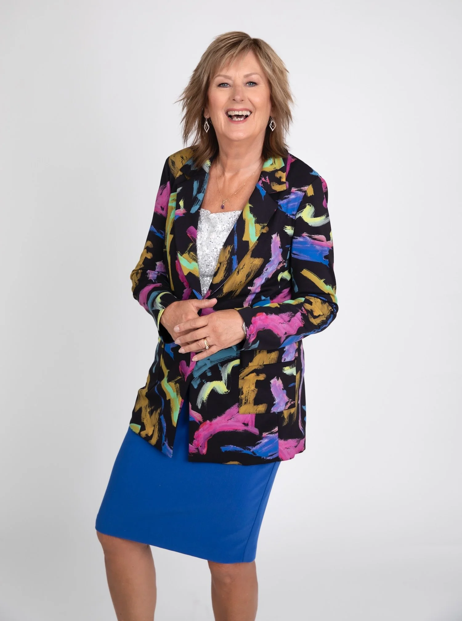 Smiling woman with short light brown hair wearing a colorful abstract patterned blazer, white blouse, and bright blue skirt, standing against a plain light gray background.