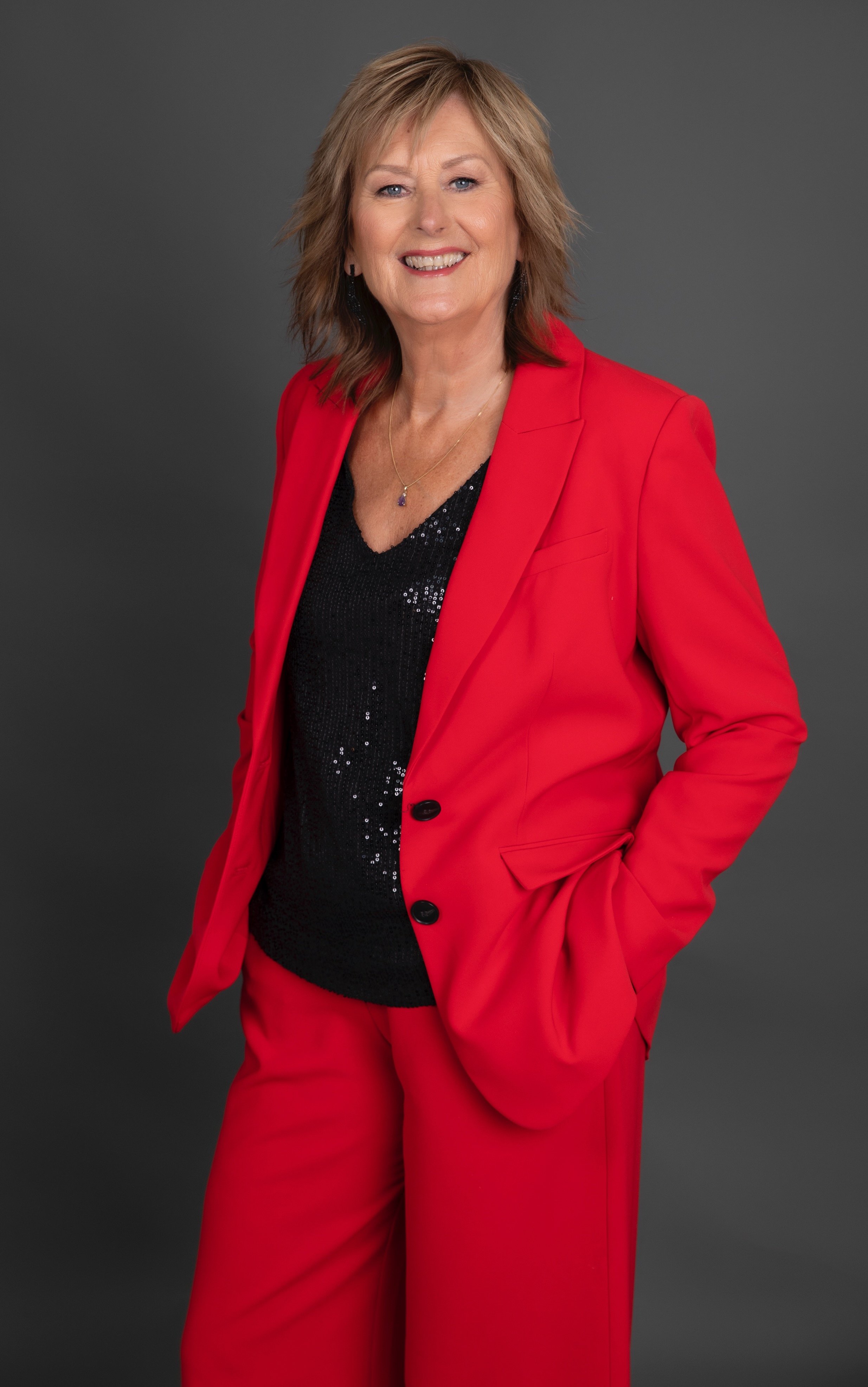Portrait of a smiling middle-aged woman with light brown hair, wearing a red suit with a black sequined top, against a gray background.