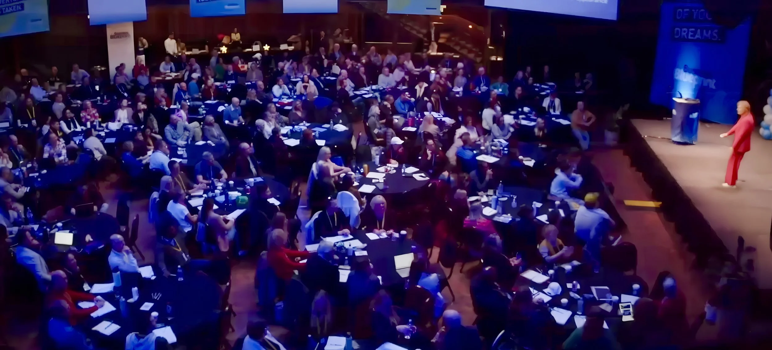 A large conference hall filled with people seated at round tables, listening to a speaker on stage. The stage has a podium and large screens displaying text.
