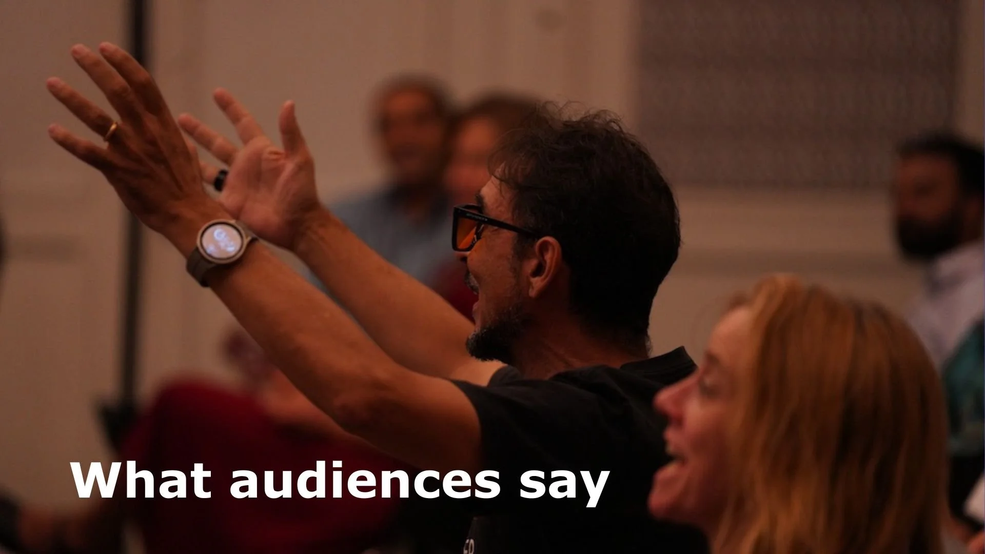 Man with glasses and beard raising his hands in applause at a conference, with woman with red hair seated nearby, and other attendees in the background.