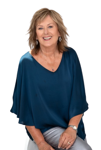 A smiling woman with shoulder-length blonde hair, wearing a blue top and silver jewelry, sitting and looking at the camera.