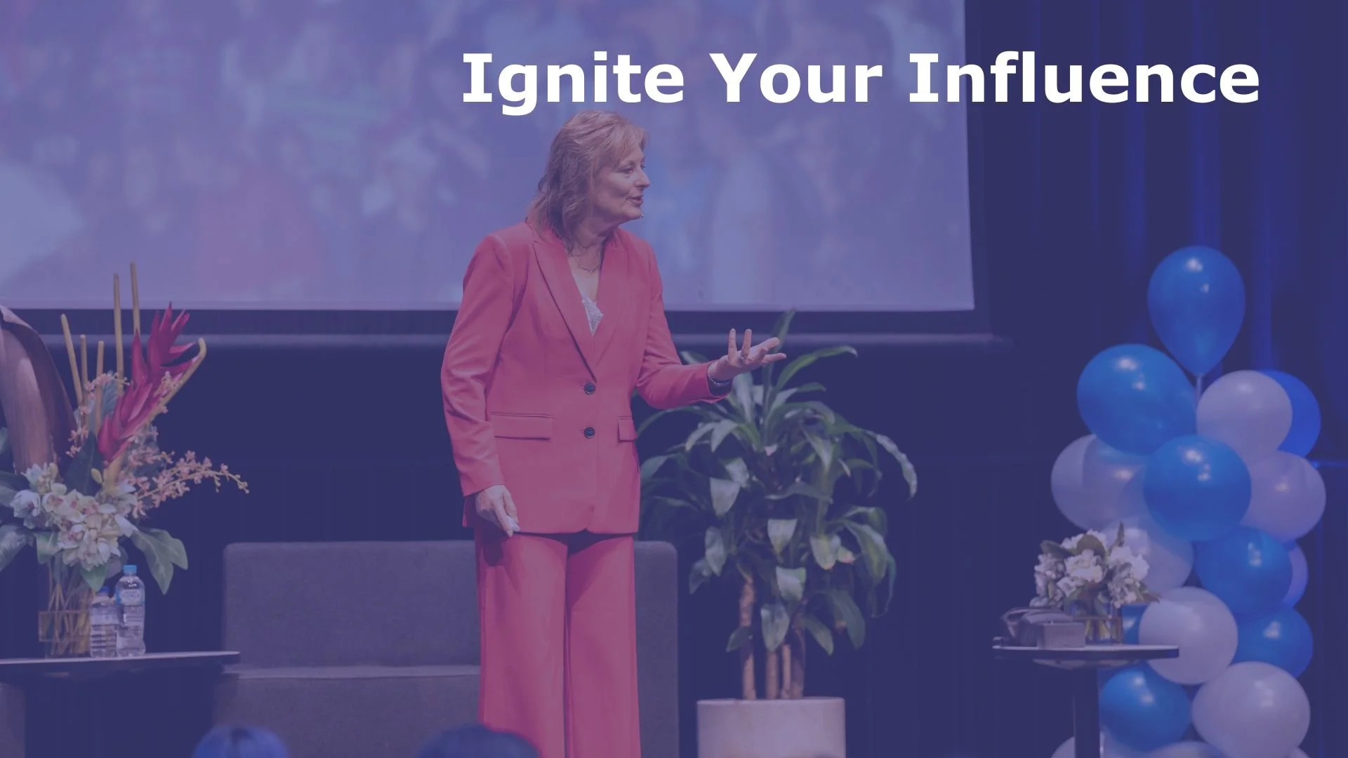 Dr Neryl East stands on stage, gesturing while speaking at an event with a large screen behind her and blue and white balloons to her right.