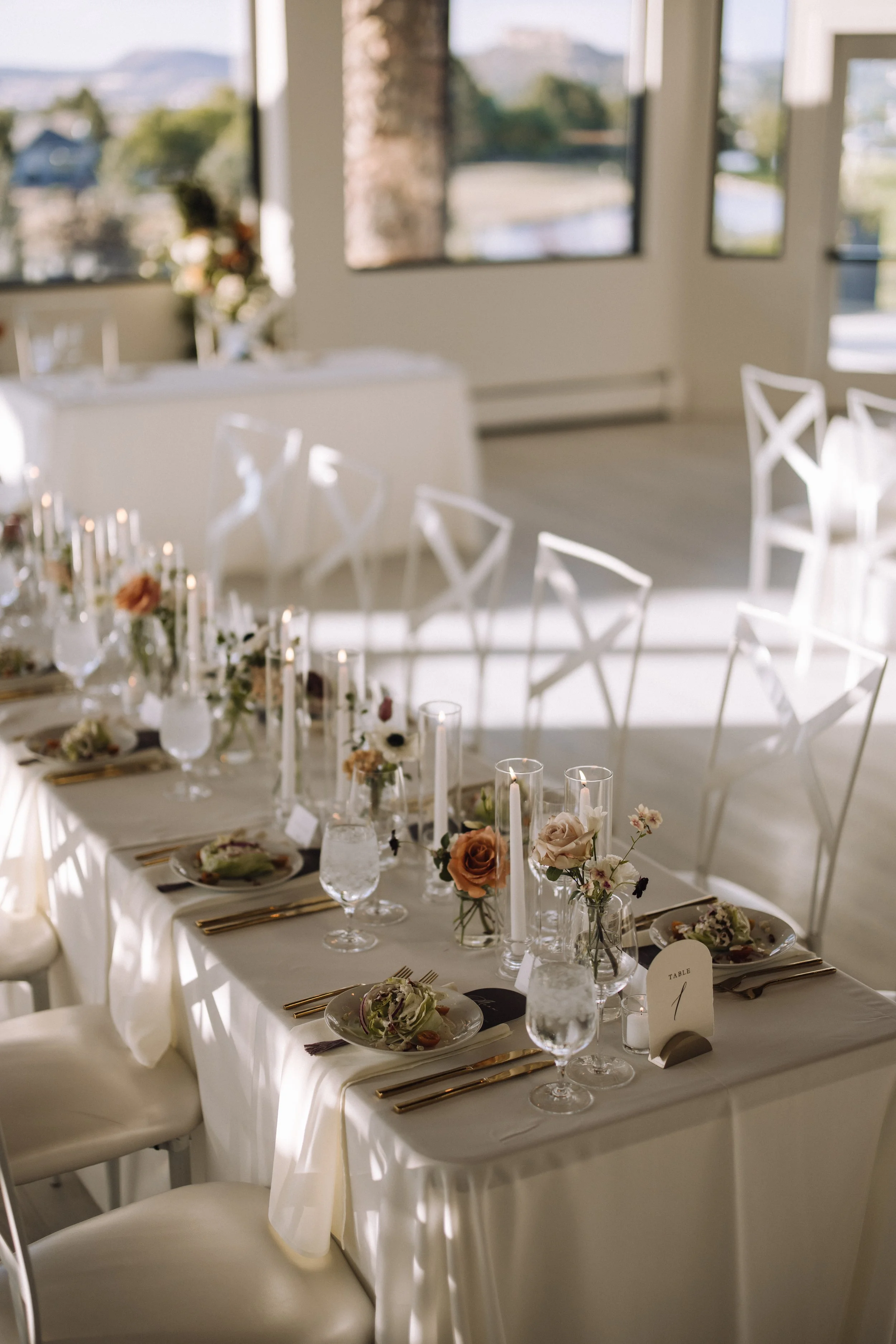 Wedding reception tablescape featuring delicate glass bud vases with peach and mauve roses, white taper candles, and gold flatware in the ballroom at The Oaks at Plum Creek in Castle Rock, CO by Grace in Bloom Floral Design.
