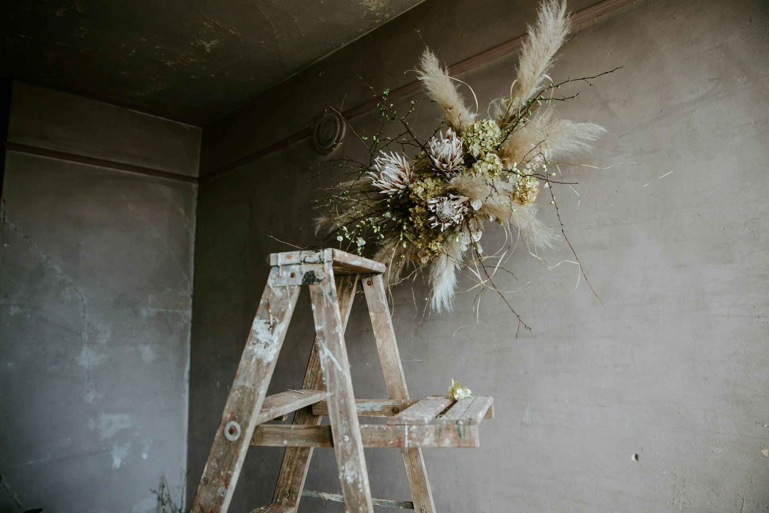 A bouquet of dried flowers and grasses arranged on a wooden stairs against a neutral gray wall and ceiling.