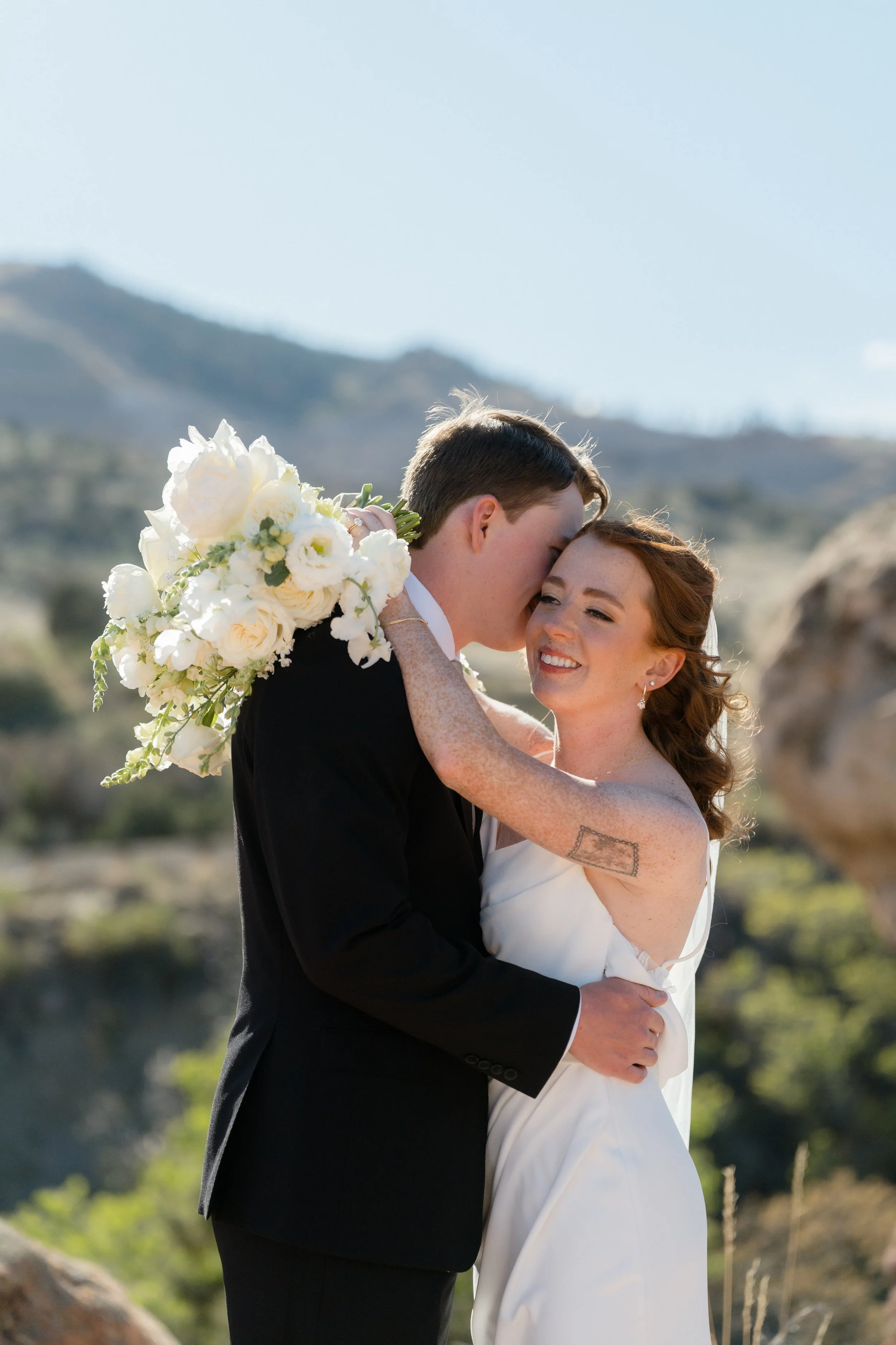 A couple in wedding attire embracing outdoors, with the man holding a bouquet of white flowers, set against a mountainous landscape with clear blue sky.