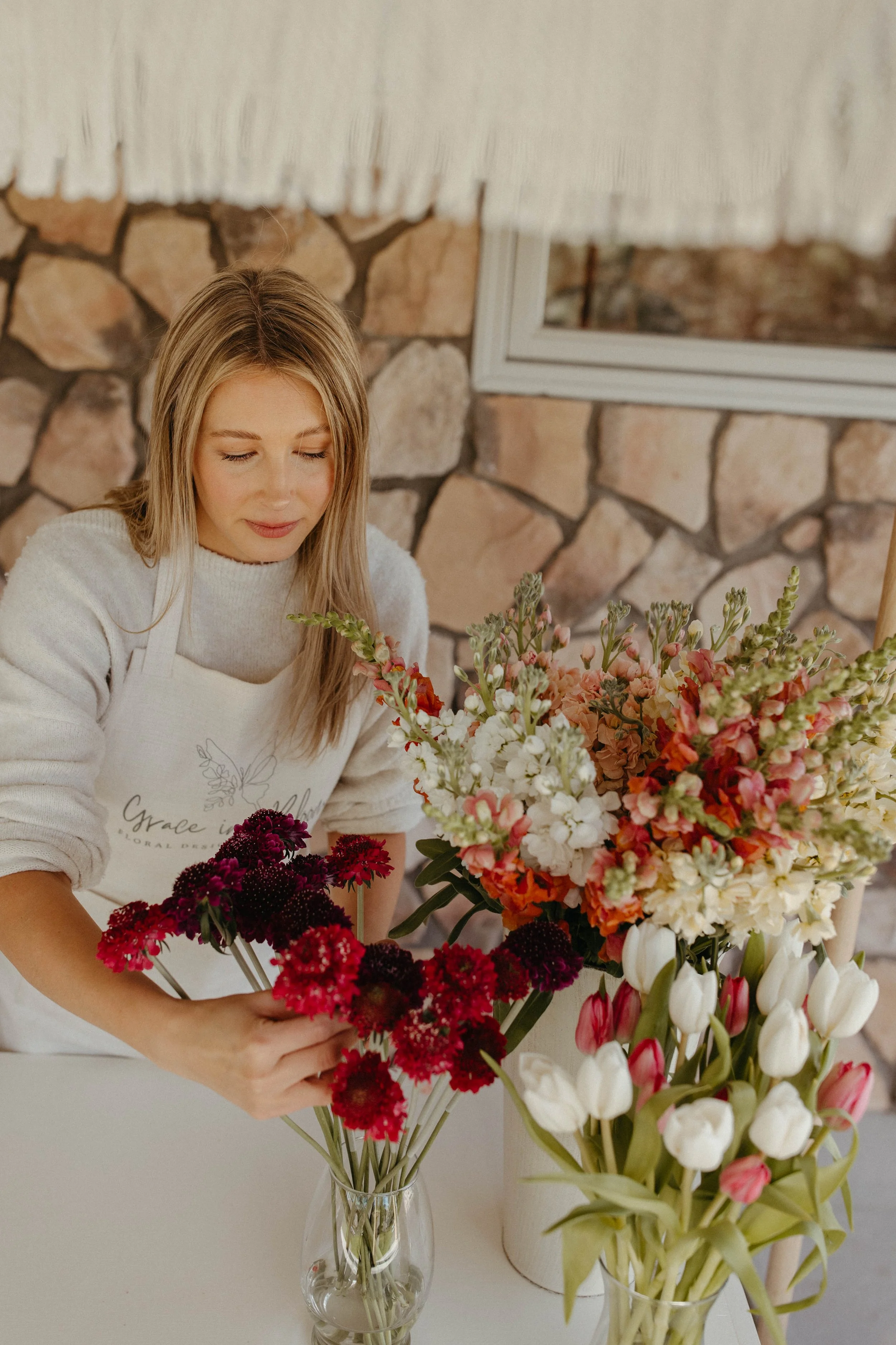 A woman arranging a bouquet of flowers at a table with other floral arrangements, with a stone wall and window in the background.