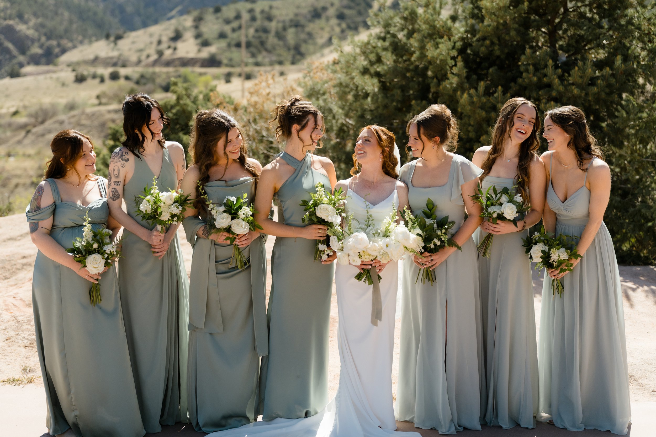 A group of nine women dressed in matching sage green and white dresses, standing outdoors in a natural setting with mountains and trees, smiling and holding bouquets of white flowers, at a celebration or wedding.